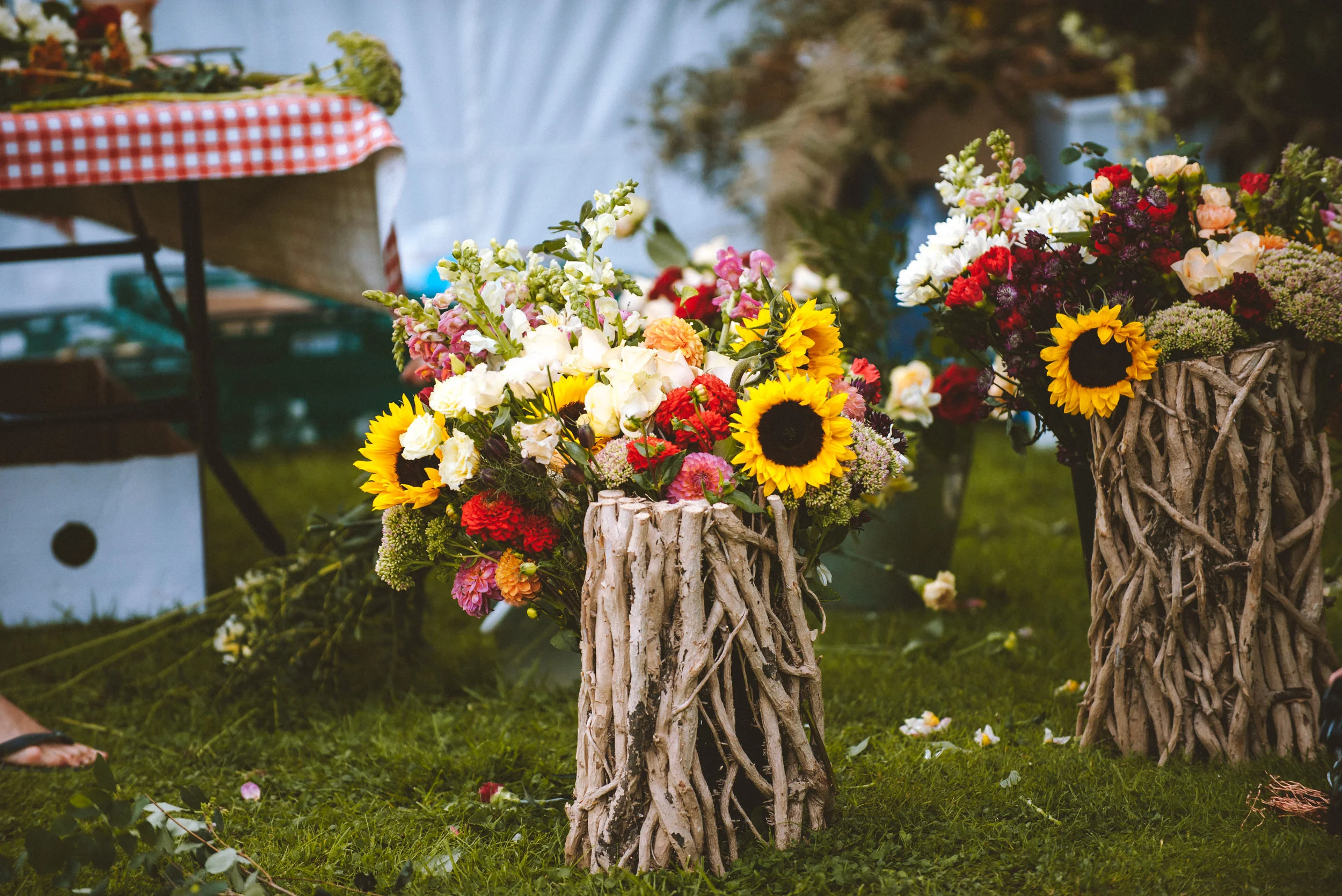 Colorful bouquet of flowers, including sunflowers, arranged in driftwood vase on green grass at an outdoor event.