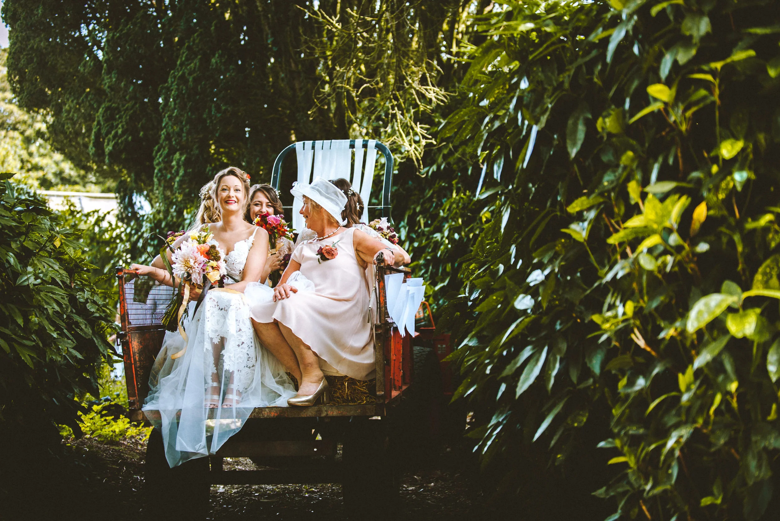 Multiple women, including a bride in a wedding dress, sitting on a vintage green wagon in a lush garden, smiling and holding bouquets of colorful flowers.