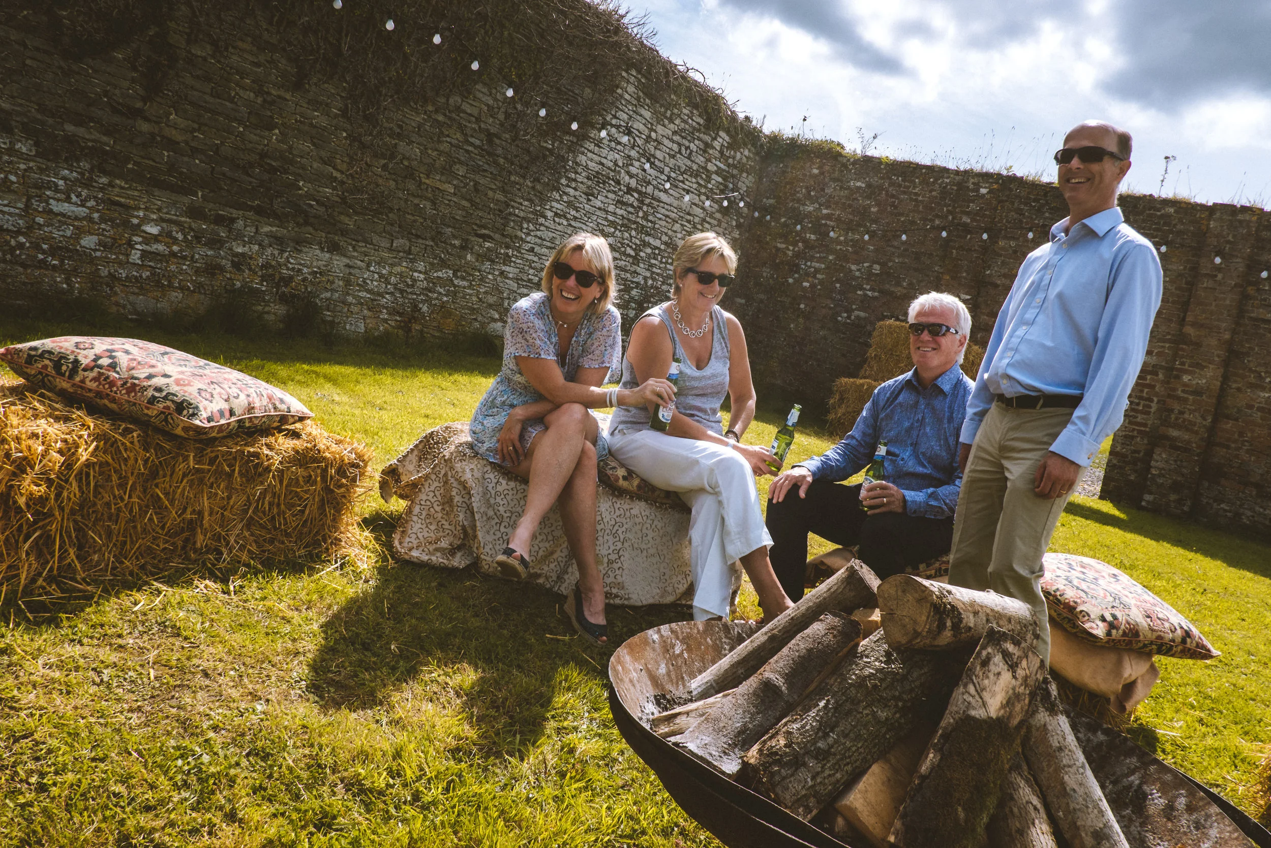 Four adults sitting and standing around a fire pit in a grassy backyard, smiling and holding drinks, with hay bales and pillows for seating, and a brick wall in the background on a partly cloudy day.