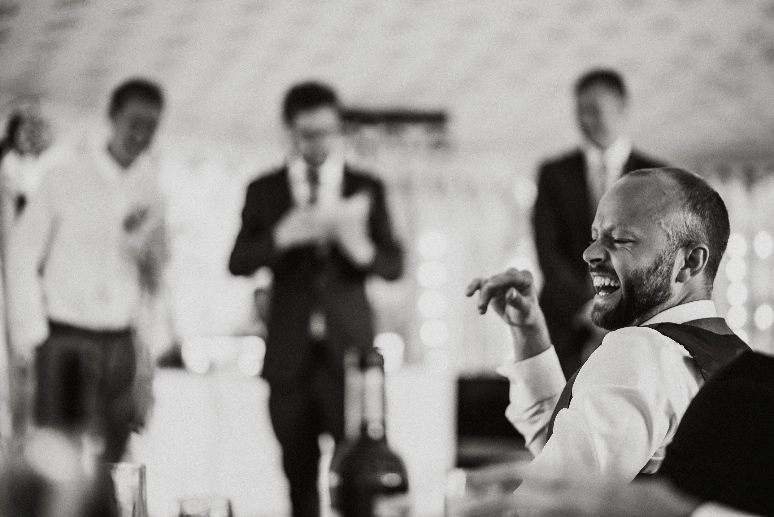 A man with a beard laughing and enjoying himself at a social gathering, with several people in the background, some reading and some standing, blurred in a black-and-white photo.
