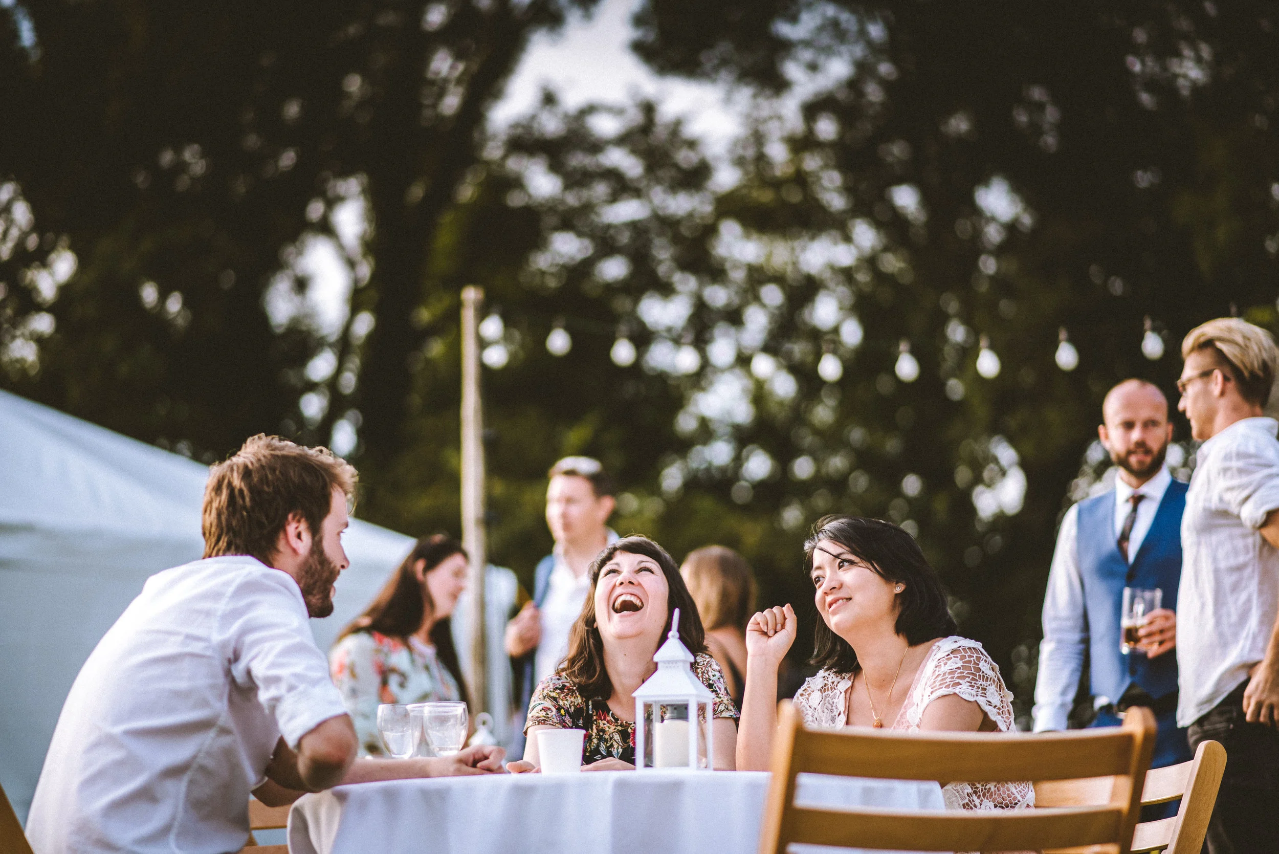 People enjoying an outdoor gathering, sitting at a table with drinks and a lantern, under string lights and trees, smiling and engaging in conversation.