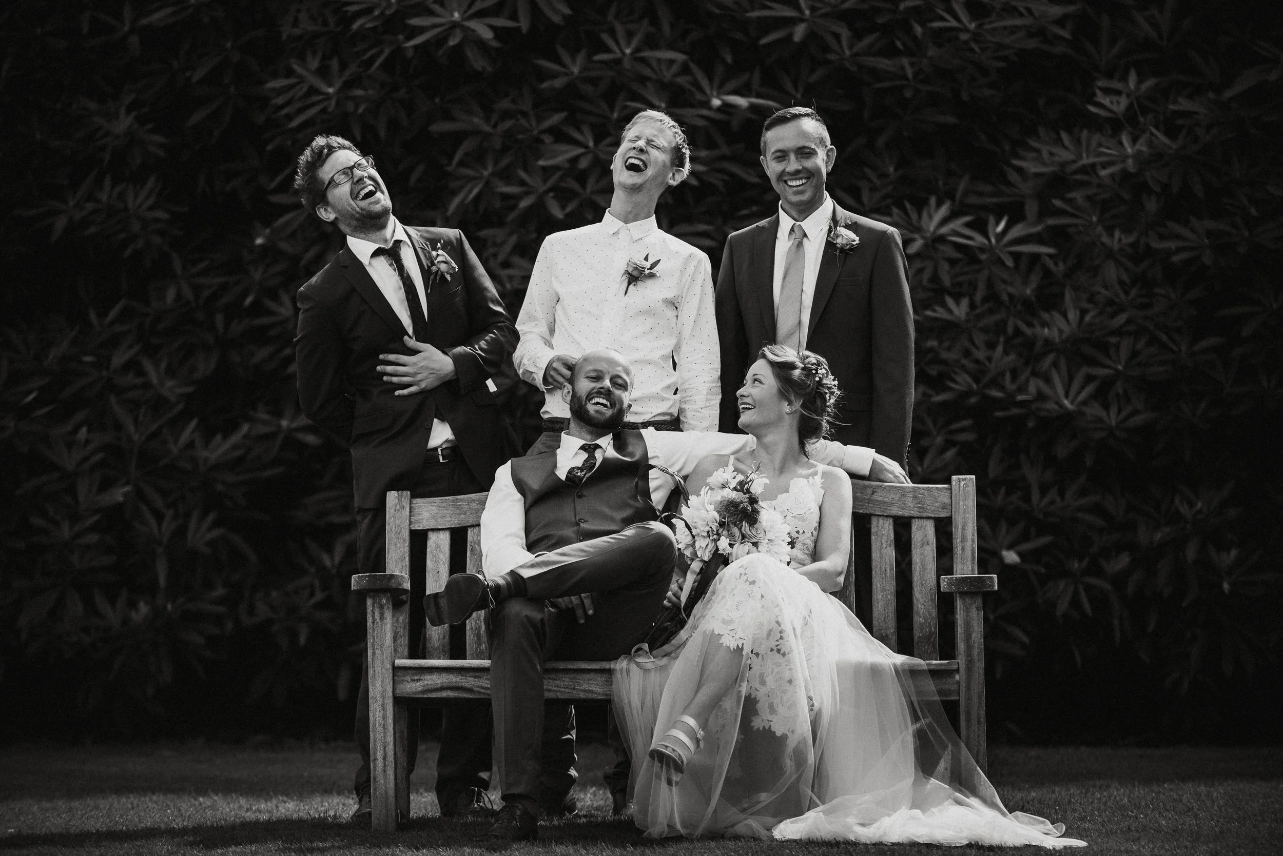 Black and white photo of a wedding party outdoors, with six people. Four men are standing, two are sitting on a wooden bench, and a woman in a wedding dress is sitting on the bench holding a bouquet. They are all laughing and appear to be very happy.