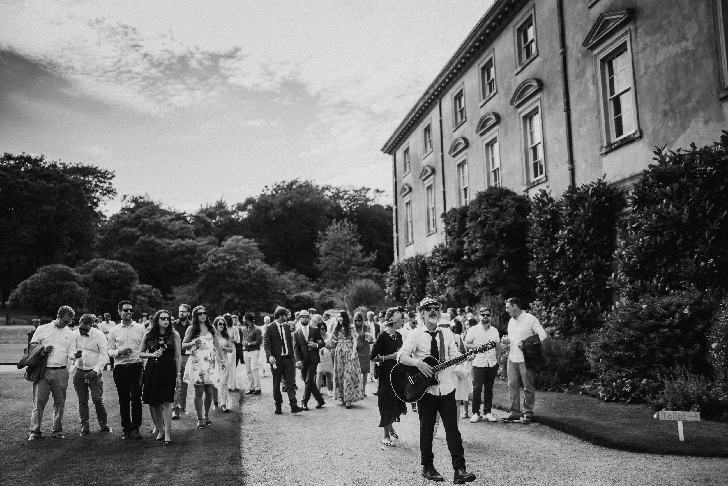 A group of people walking on a gravel pathway, some are wearing sunglasses, and a man in the front is playing a guitar and singing. The scene is outdoors beside a large historic building with trees in the background and a sign pointing to the toilets