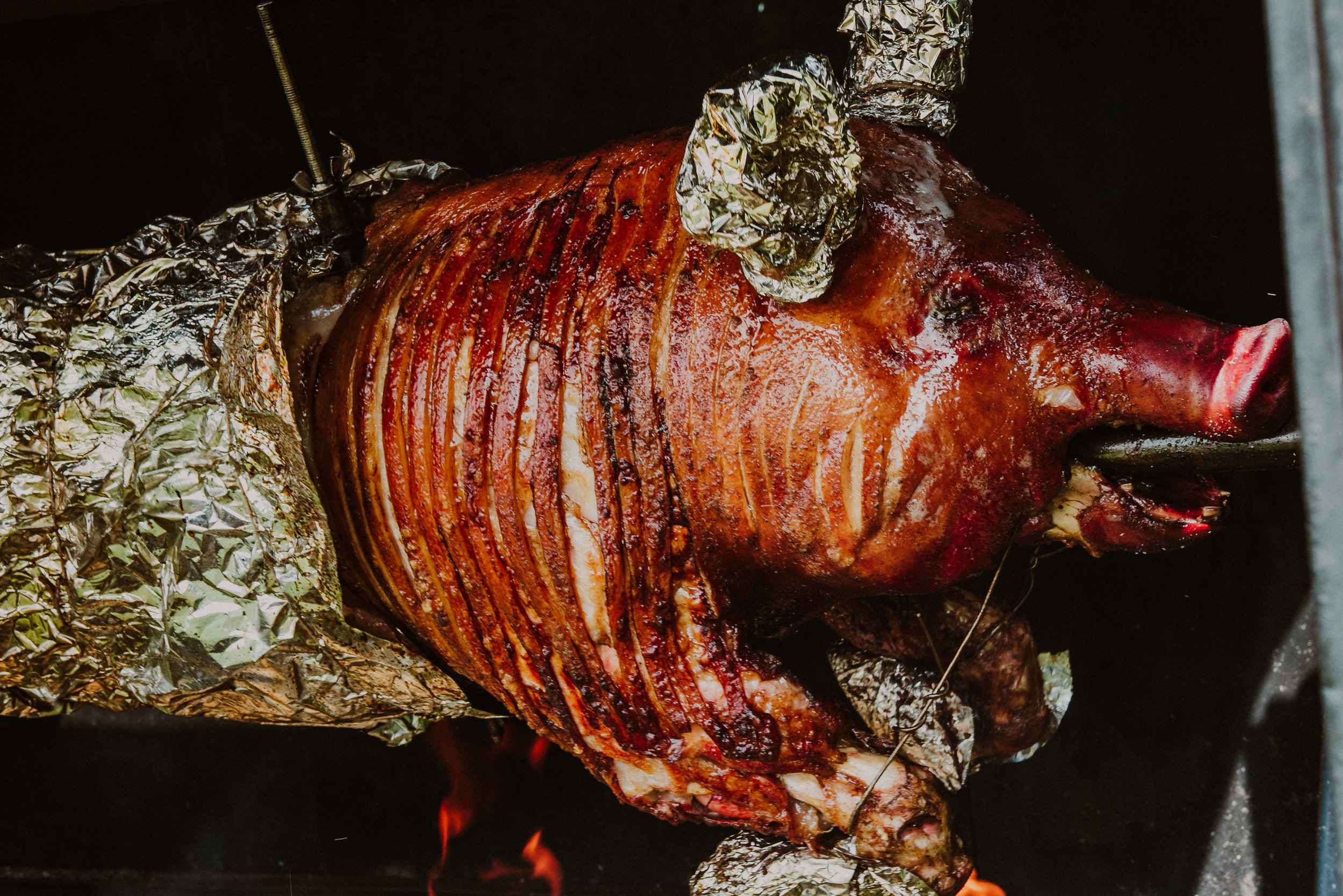 A whole roast pig with crispy skin, wrapped in aluminum foil at the tail, cooking in an oven or pit fire.