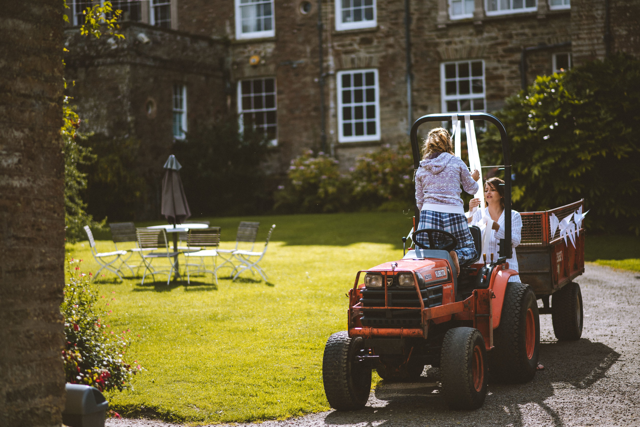 Two girls playing on a red tractor with a trailer in a garden with green grass and a brick house in the background.
