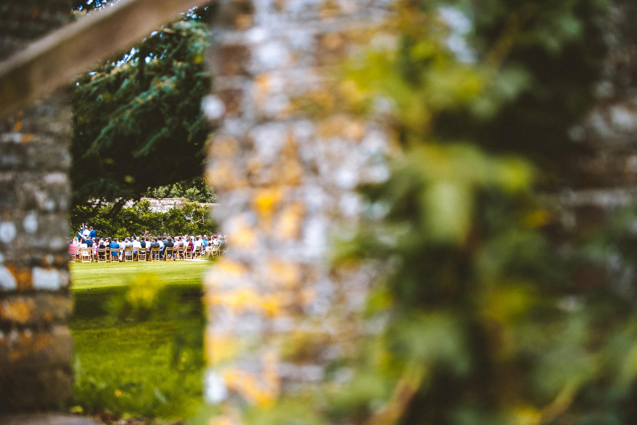 A group of people sitting at tables in a garden, seen through a gap in a stone or brick wall.