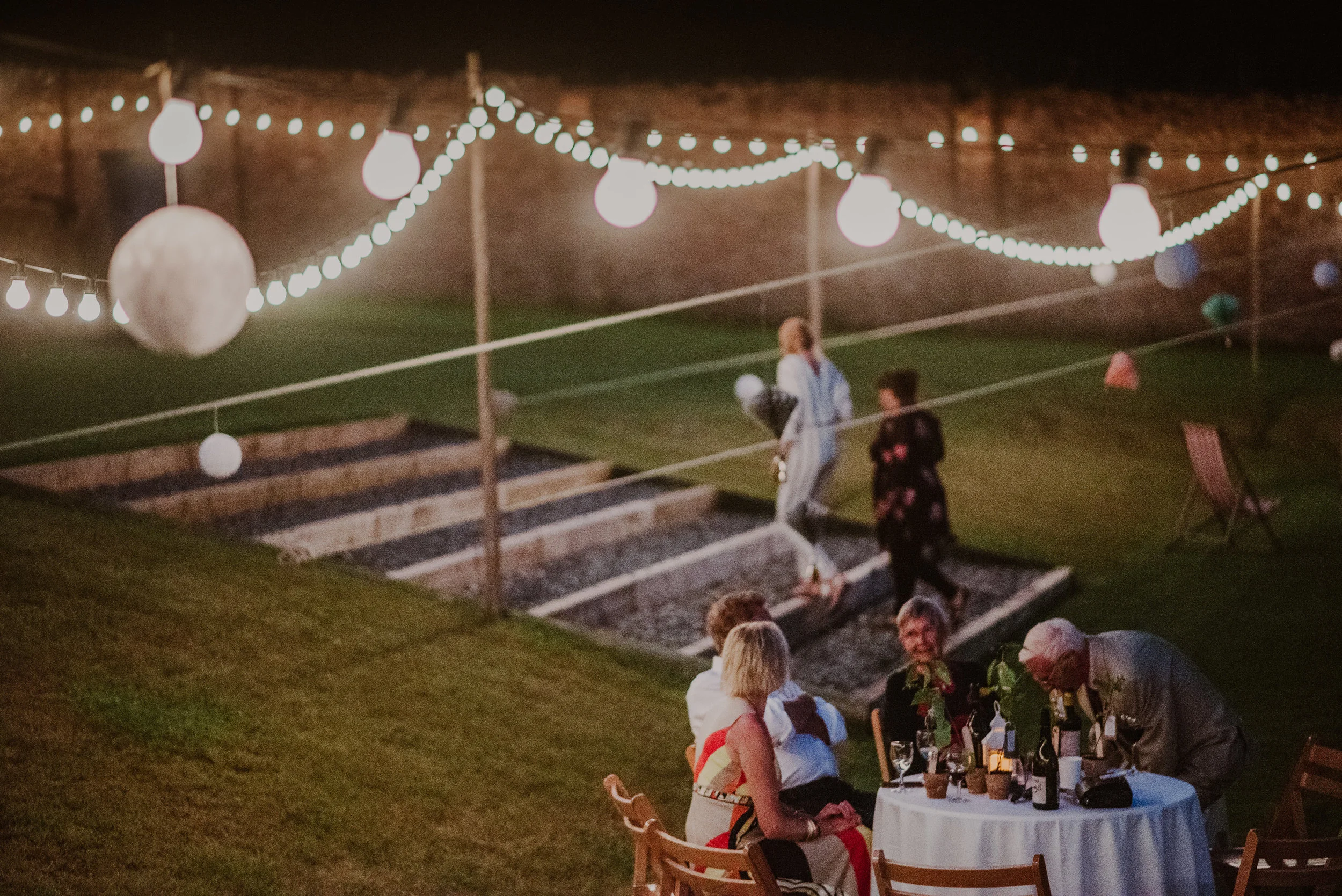 People dining outdoors at a round table with wine bottles and glasses, decorated with candles and flowers, under string lights with paper lanterns, on a grassy lawn at dusk.