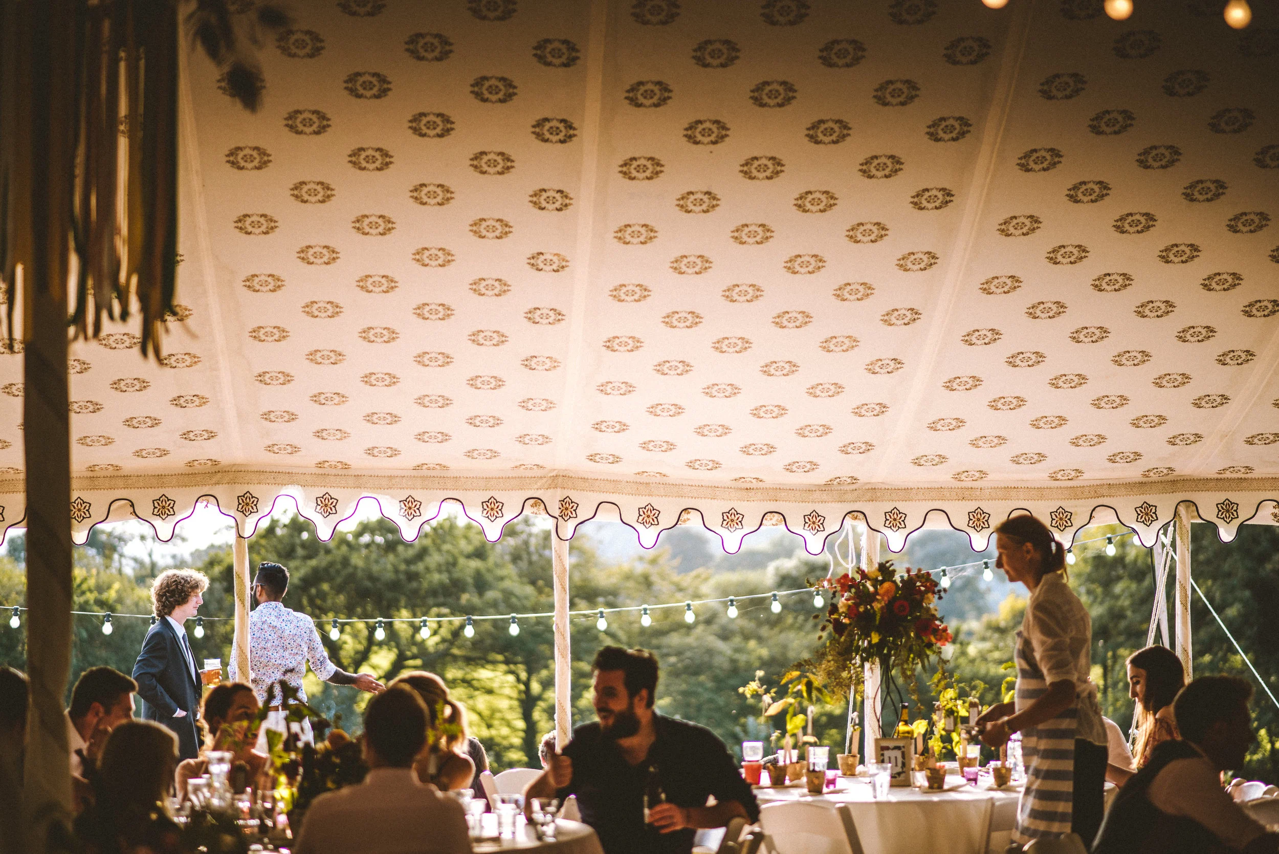 People gathered at a sit-down event under a large tent outdoors, with trees in the background. Some individuals are sitting while others stand, and a woman is serving drinks. The tent has a decorated ceiling and fairy lights outside.