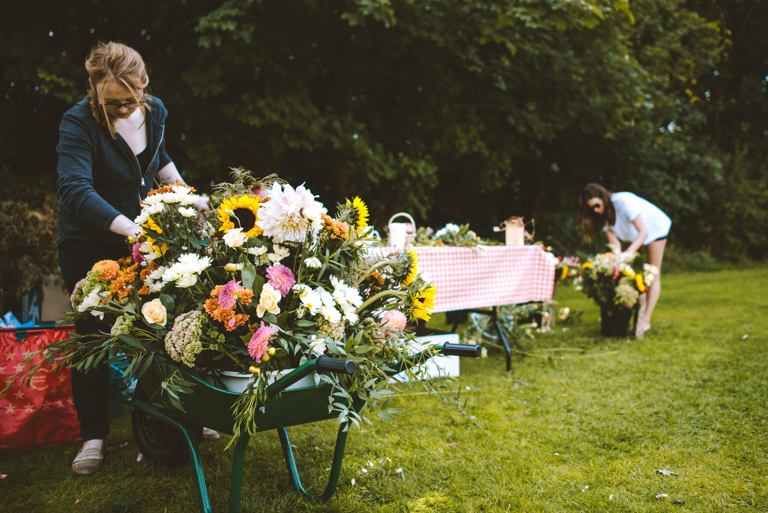 Two women preparing flower arrangements outdoors on a sunny day, with a pile of colorful flowers and a table with supplies.