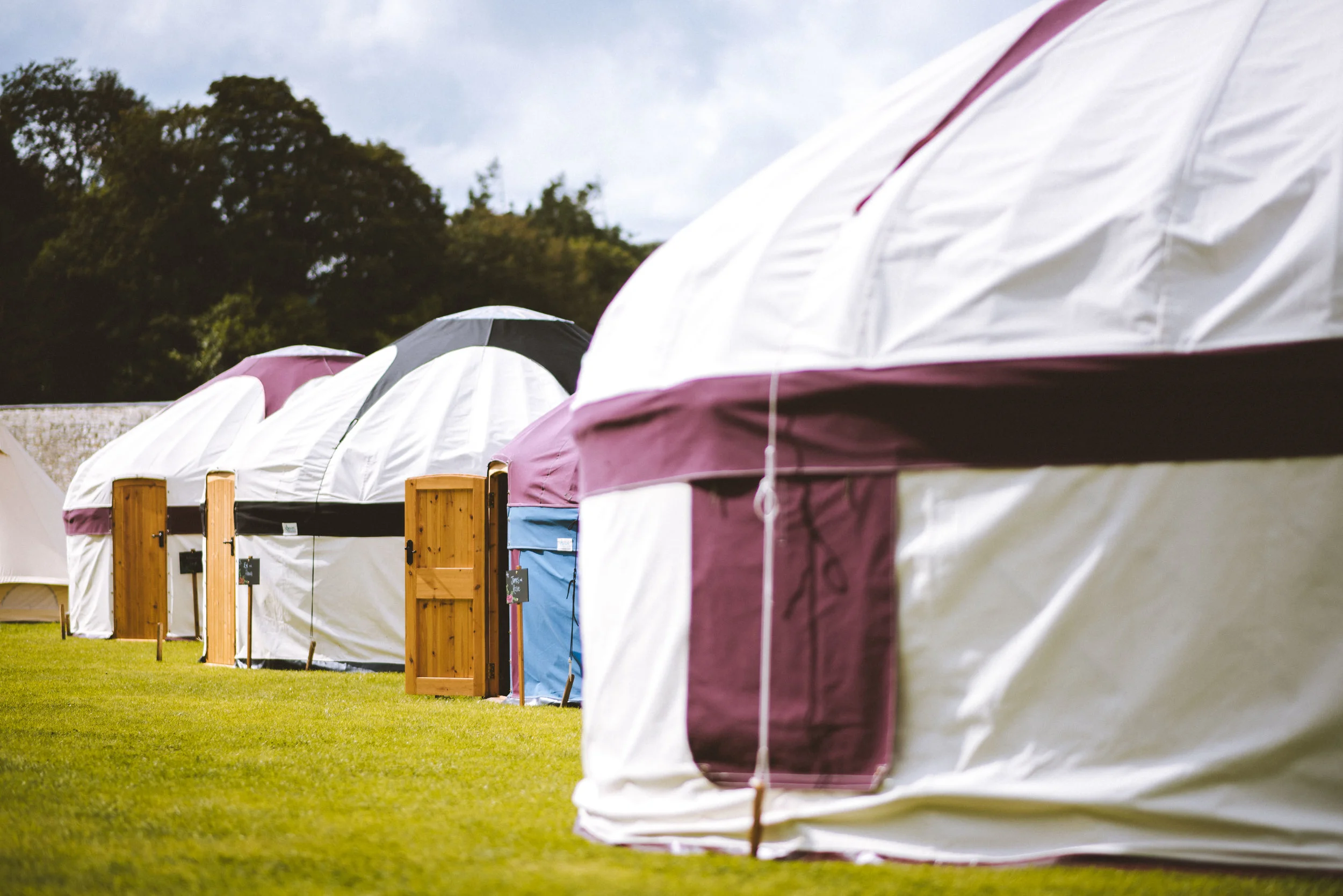 A row of white camping yurts with different colored doors and small wooden fences on a grassy field, with trees and cloudy sky in the background.