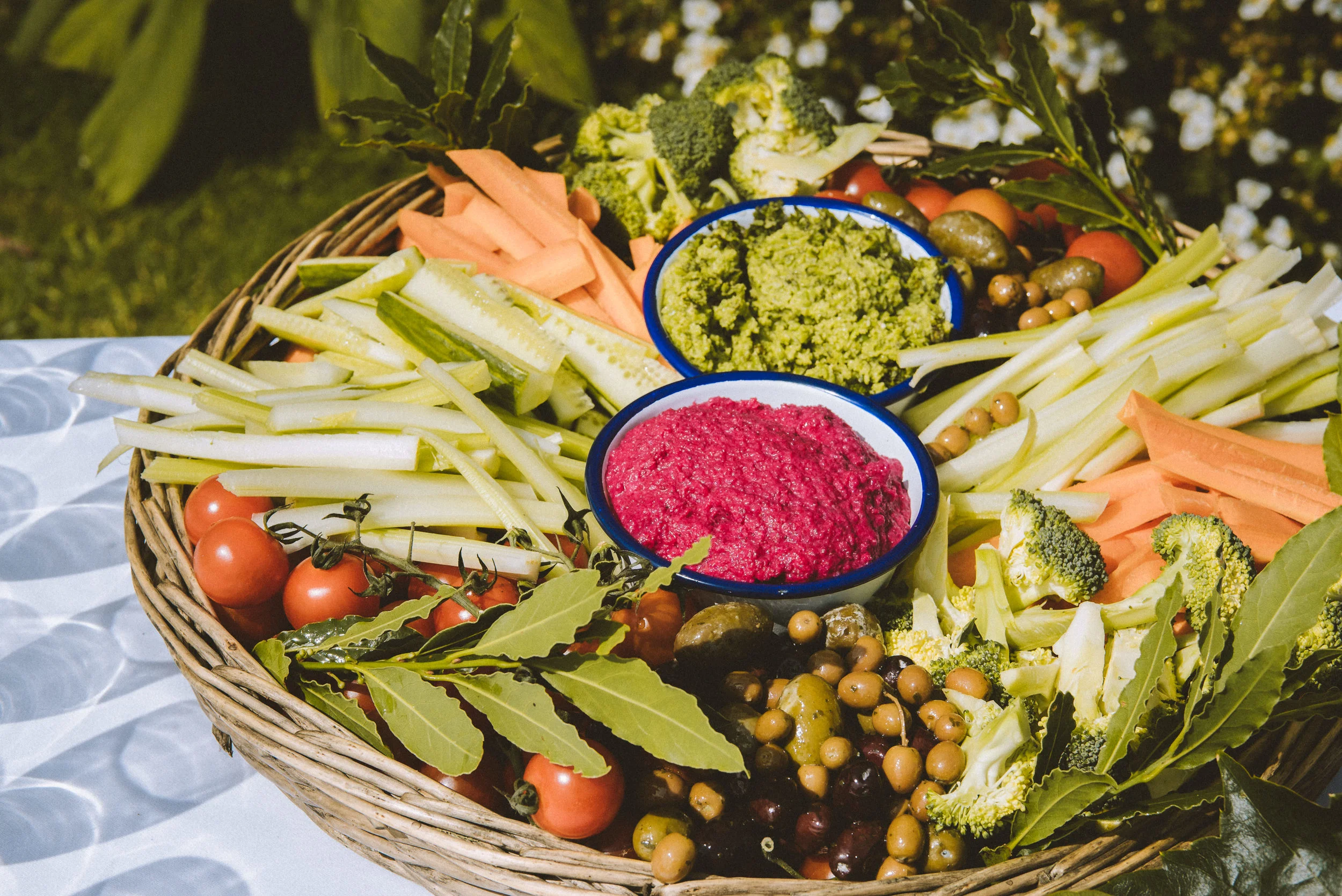 Basket filled with fresh vegetables and dips, including cherry tomatoes, sliced celery, carrots, broccoli, olives, and bowls of guacamole and beet hummus.