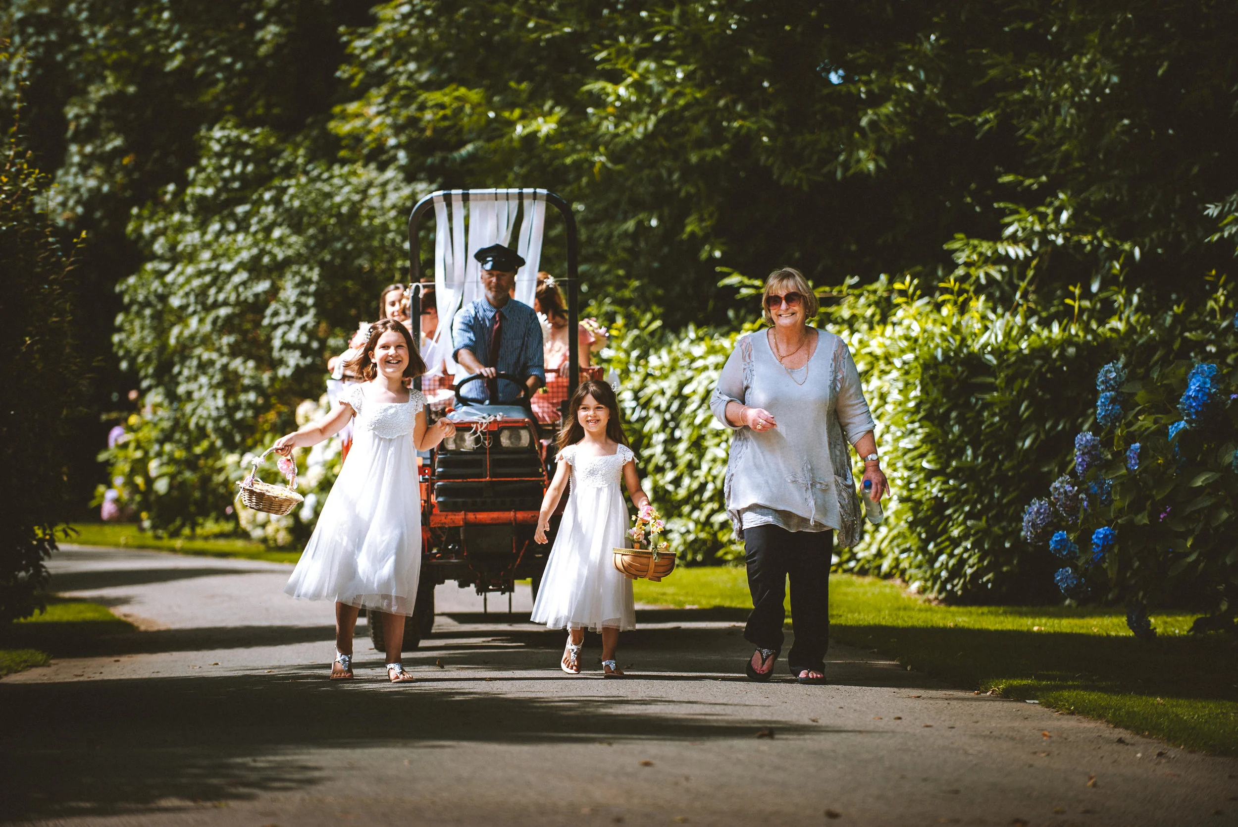 A group of children and an adult walking along a path surrounded by greenery, with some children dressed in white dresses holding baskets, and a person pushed in a stroller.