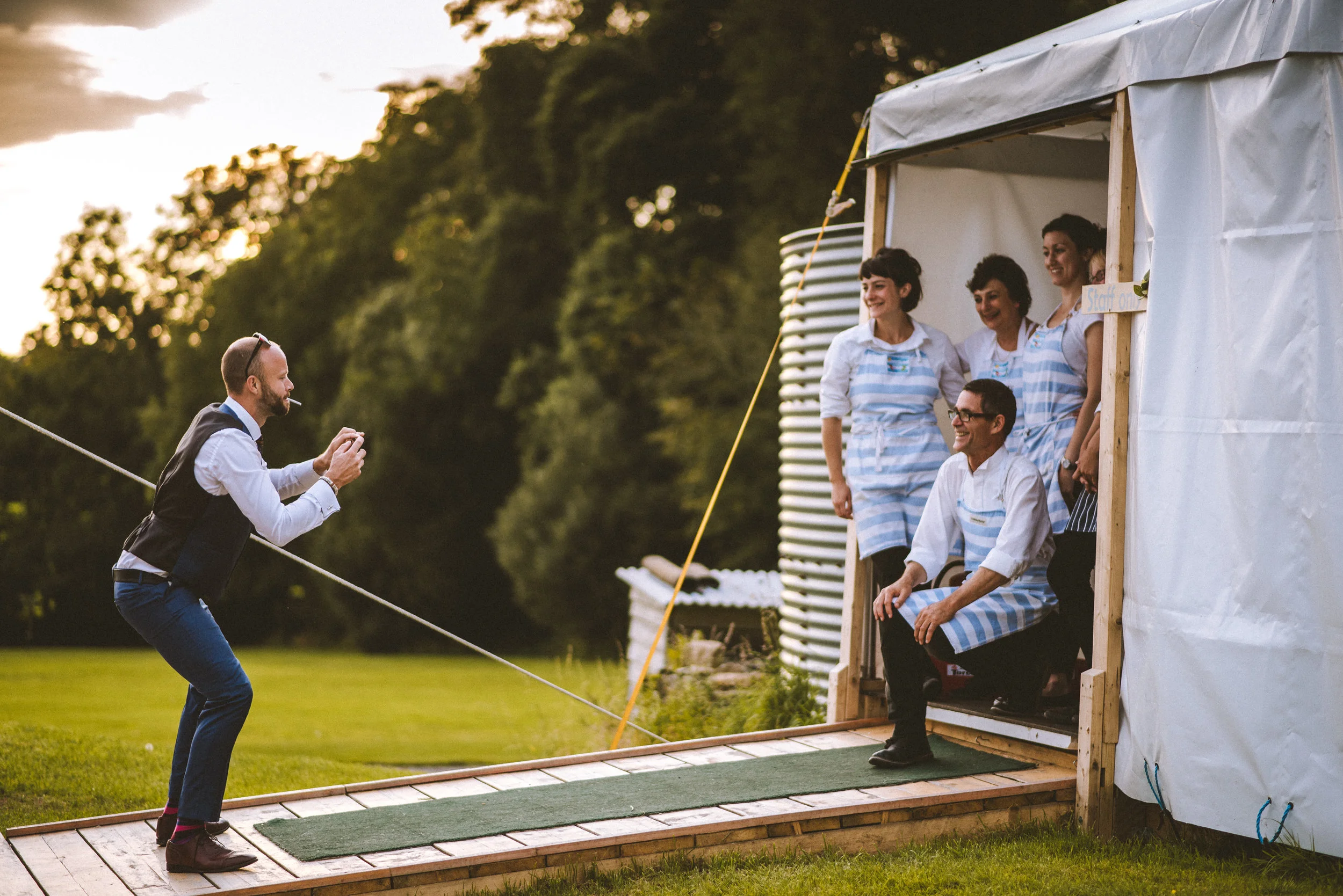 A man taking a photo with a camera or phone of four women and a man sitting on a wooden platform outside a white tent, all wearing matching blue and white striped aprons, at an outdoor event during sunset.