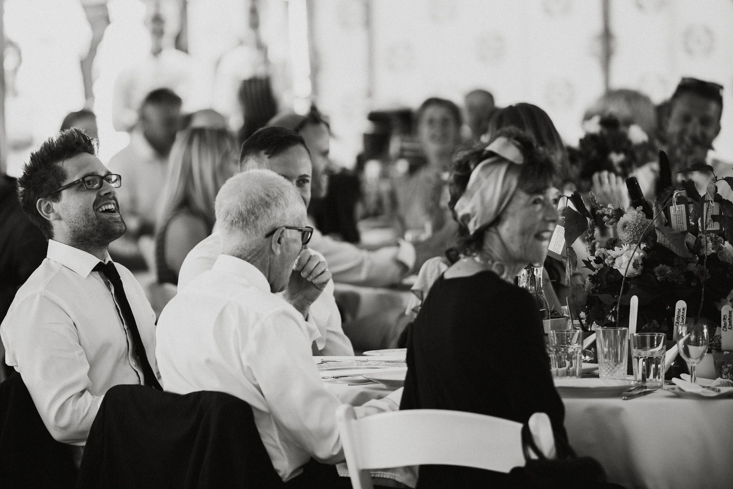 A group of people sitting at a long table, smiling and laughing during a social gathering or celebration, with floral centerpieces and glasses on the table.