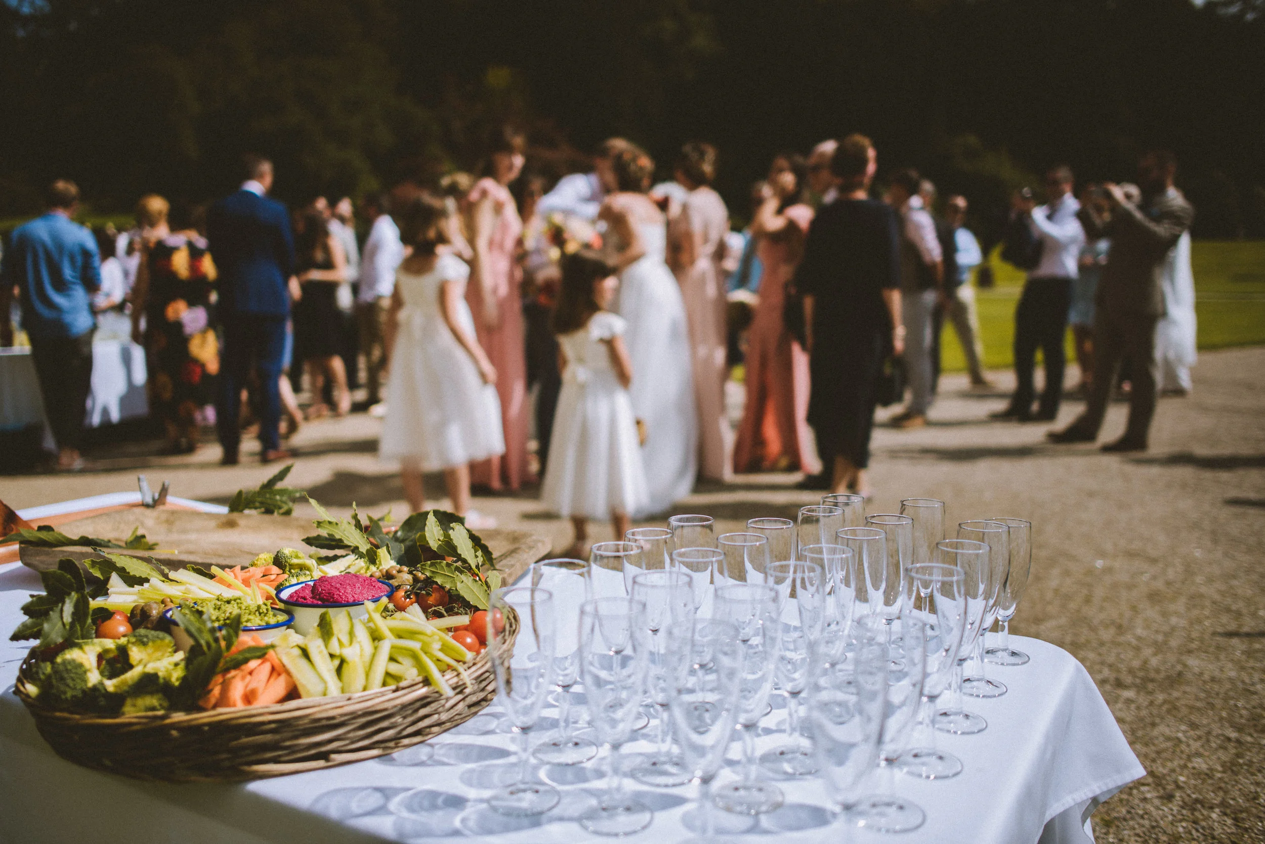 A table with empty champagne flutes and a platter of fresh vegetables, with wedding guests mingling in the background at an outdoor wedding reception.