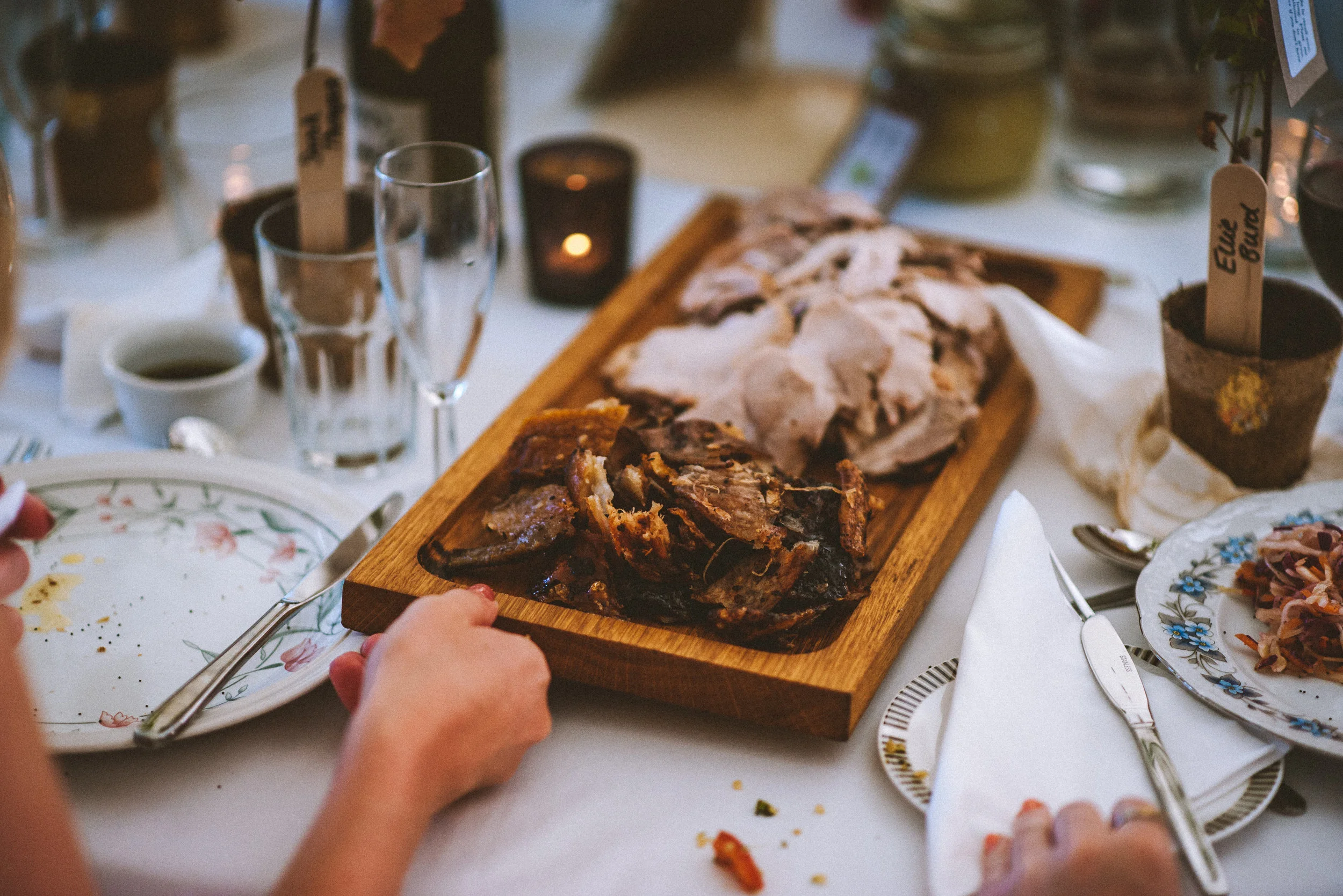 A table set for a meal with a wooden platter holding sliced cooked ham and roast beef, surrounded by plates, utensils, glasses, and small potted plants labeled 'Eucalyptus' and 'Fuchsia'.