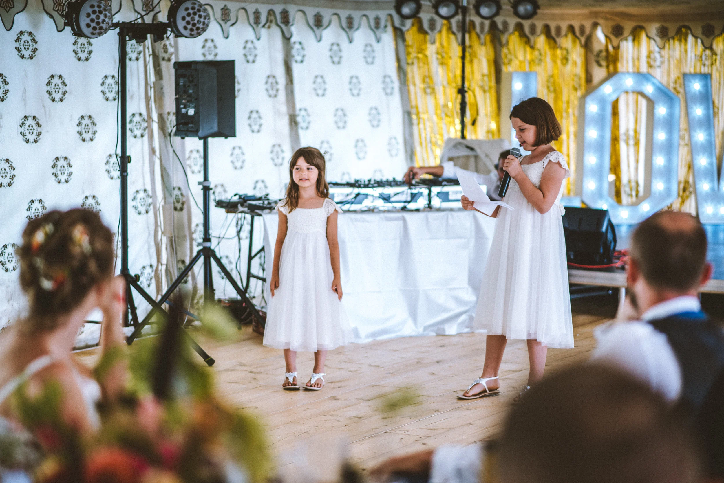 Two young girls in white dresses giving a speech at a wedding reception. One girl is reading from a paper while the other stands nearby. The background includes decorated curtains, large lighted letters, and a DJ setup. Guests are seated, observing t