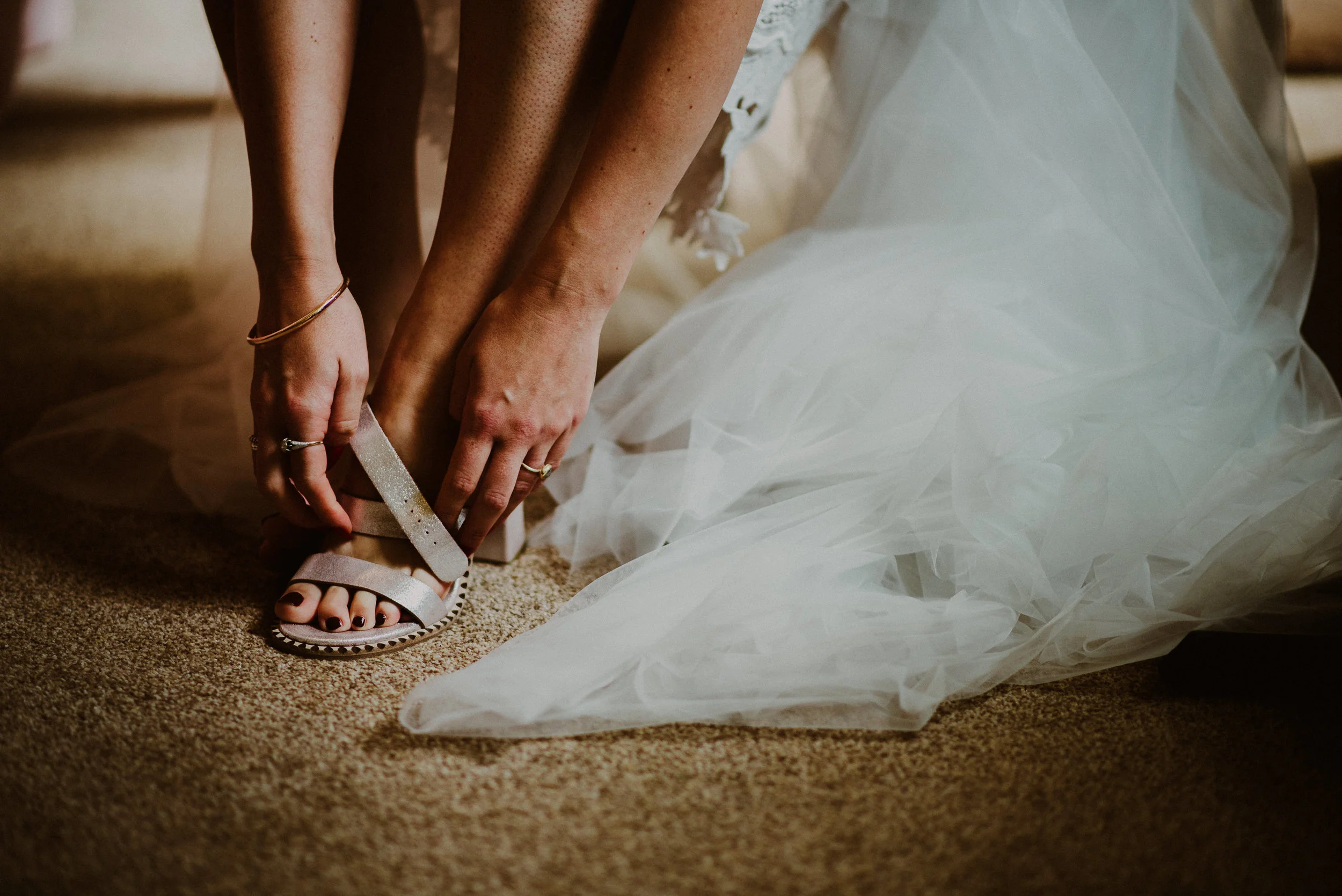 A person in a wedding dress is putting on a high-heeled sandal with glittery straps.