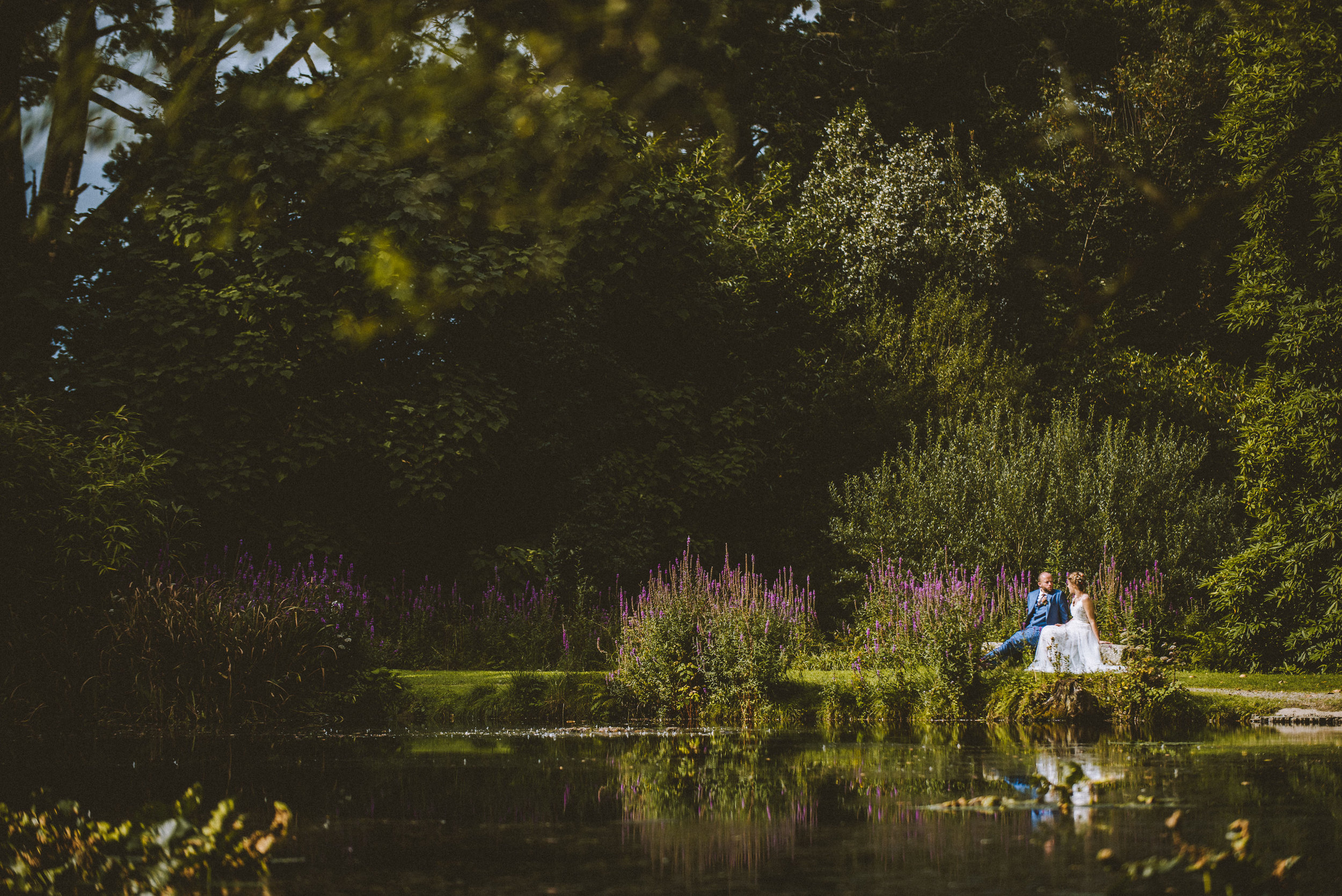 A couple dressed in wedding attire sitting on a rock by a small pond surrounded by lush greenery and purple flowers.