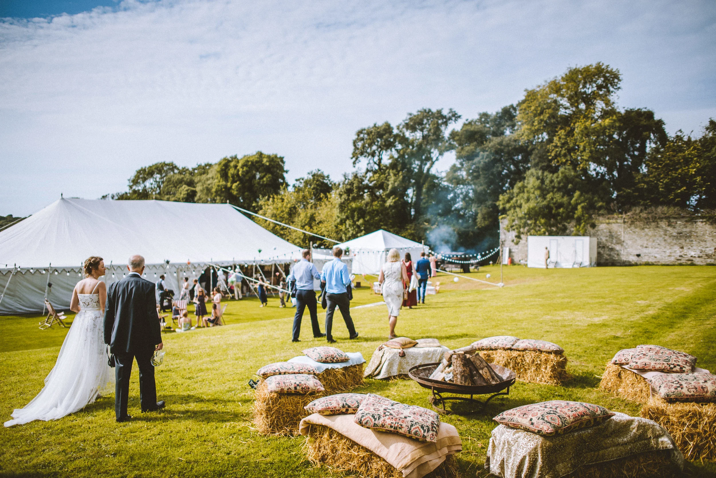 People attending a wedding reception outdoors, with a large white tent, hay bales with decorative pillows, and a grassy area with trees in the background.
