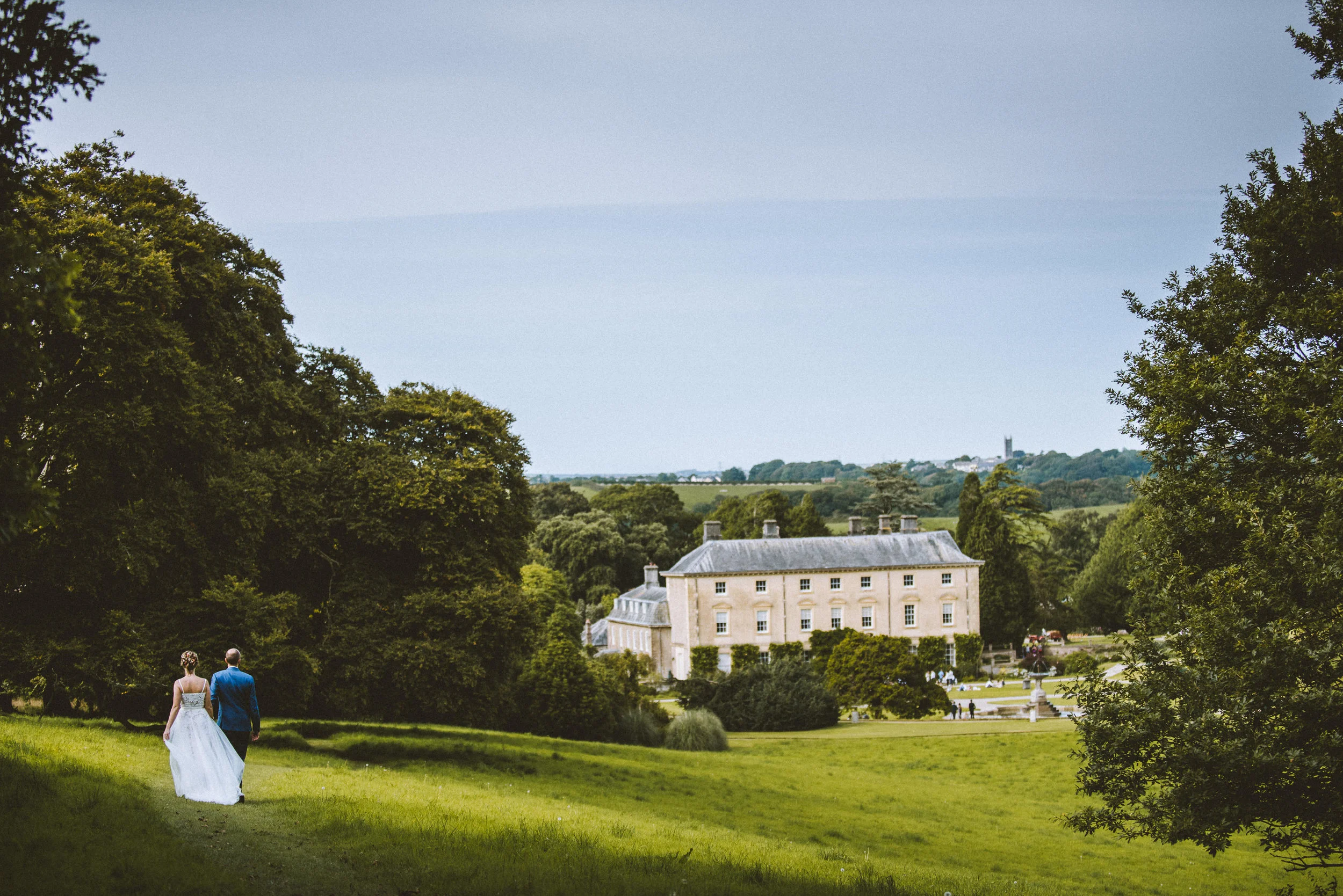 A bride and groom walking on a grassy trail towards a large, historic stone mansion surrounded by trees and gardens.