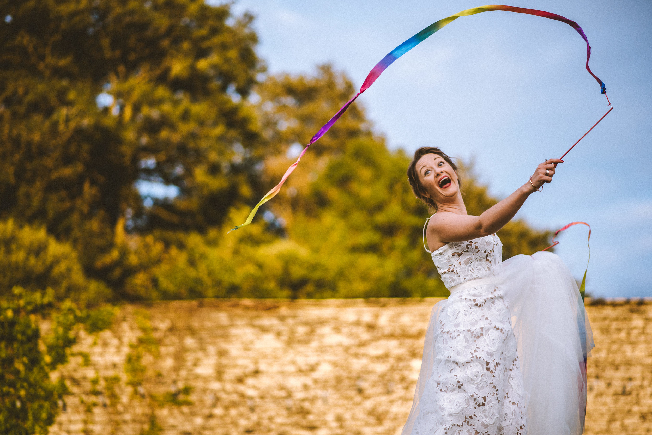 A woman in a white dress happily waving a rainbow-colored ribbon outdoor during daytime with trees and a brick wall in the background.