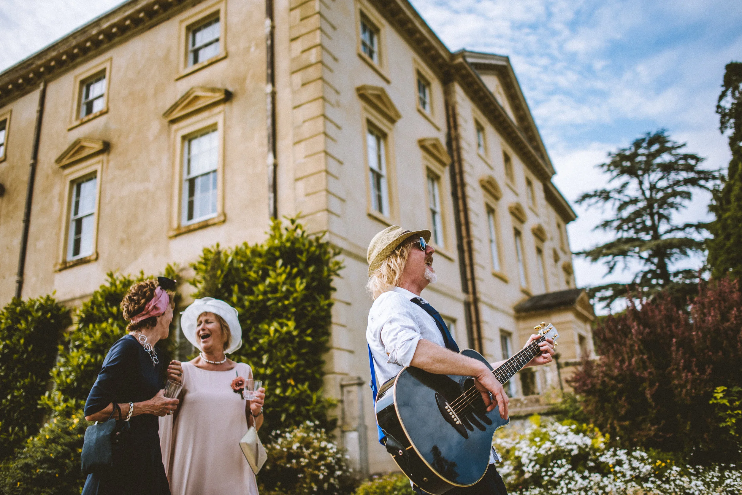 A man playing guitar outside a large, elegant building while two women in vintage dresses and hats enjoy the music and laughter.