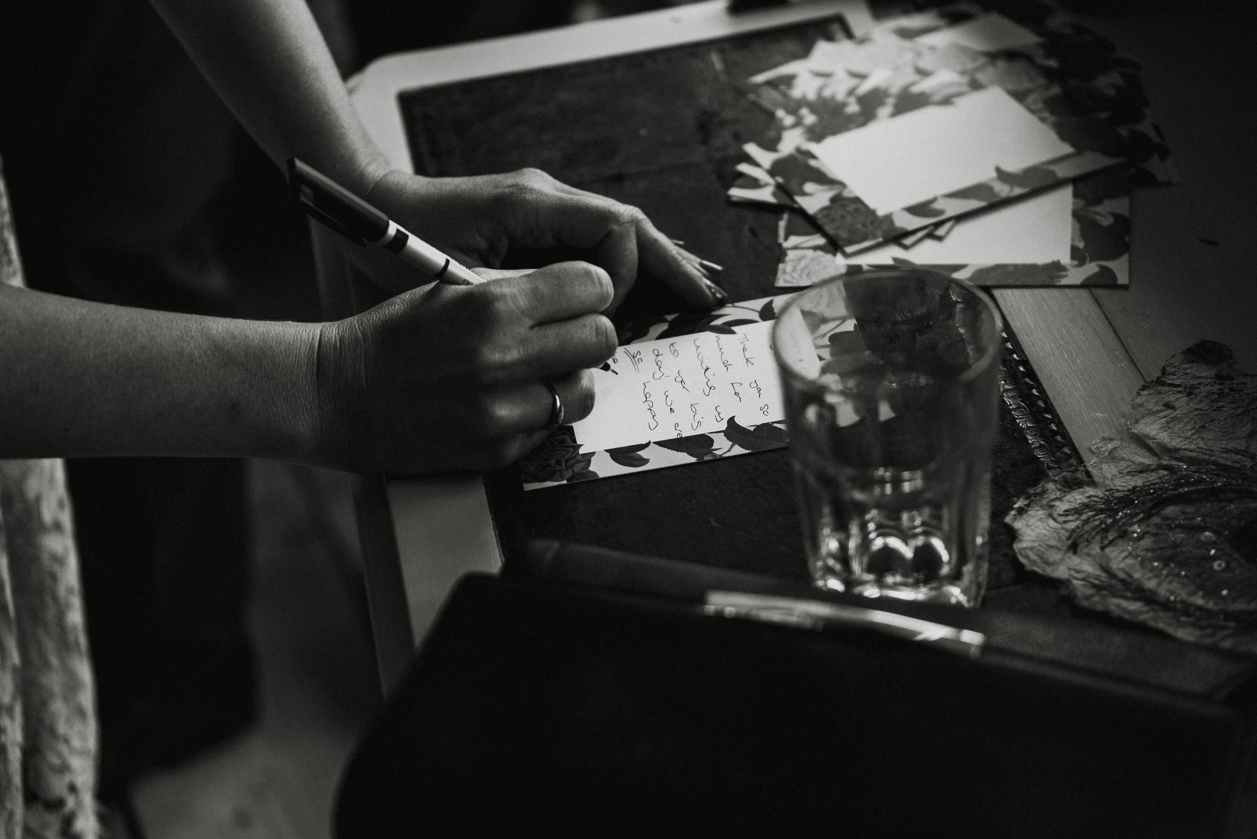 A person is writing a handwritten note on decorative paper at a table, with art supplies, a glass of water, and other papers nearby.
