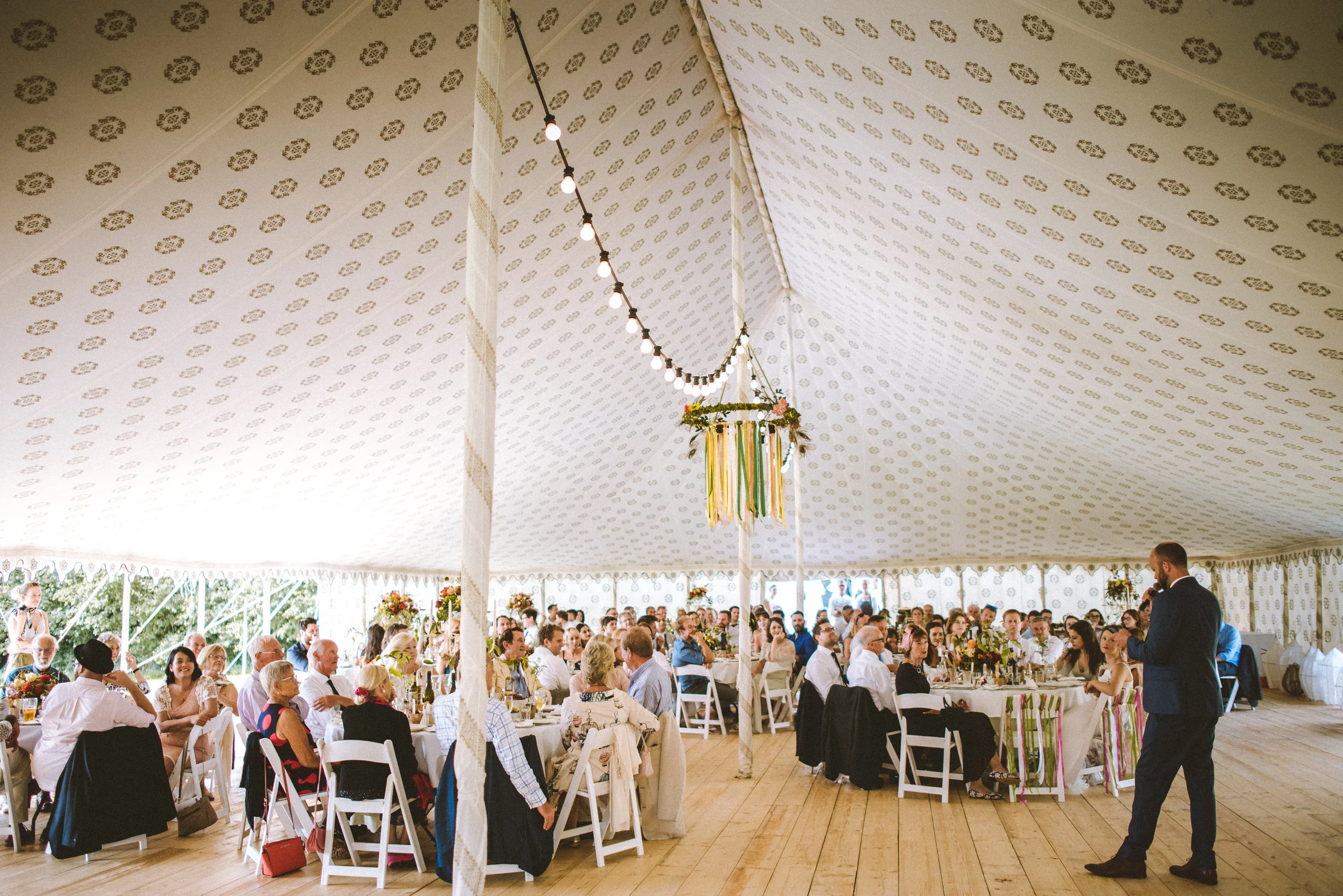 Wedding reception inside a large decorated tent with guests seated at round tables, a man in a suit giving a speech, and festive hanging decorations.