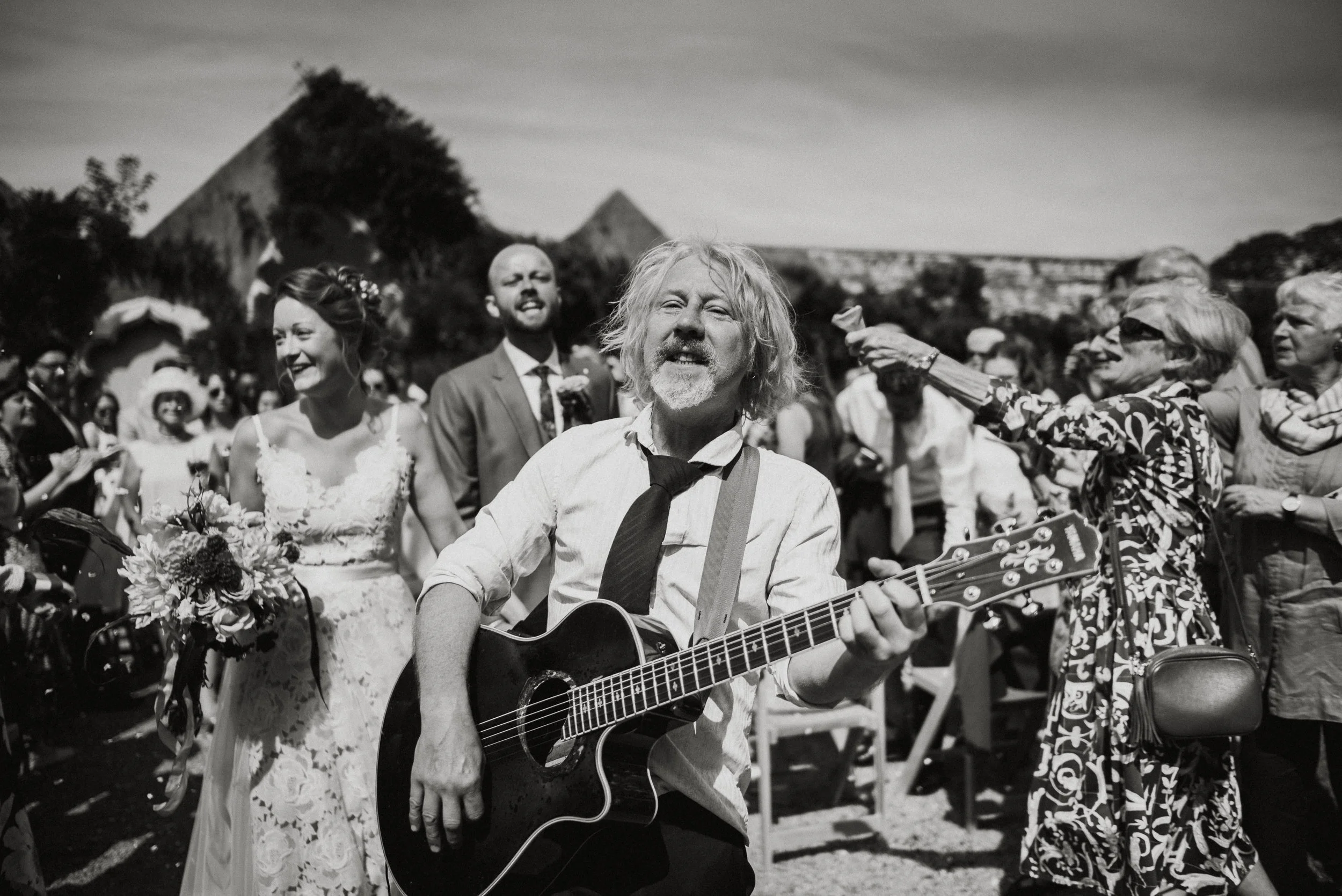 A man playing guitar outdoors at a wedding with bridesmaids and guests in the background in a black and white photo.