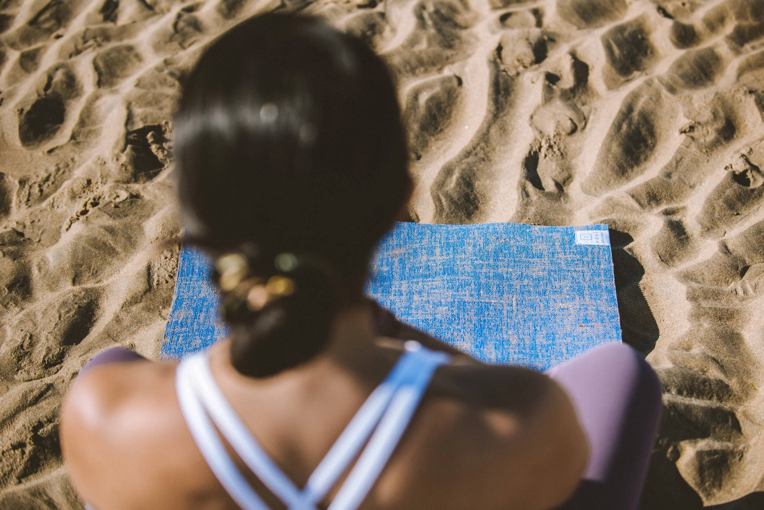 A woman sitting on a beach towel on sandy ground, with her back turned to the camera, looking towards the ocean or horizon.