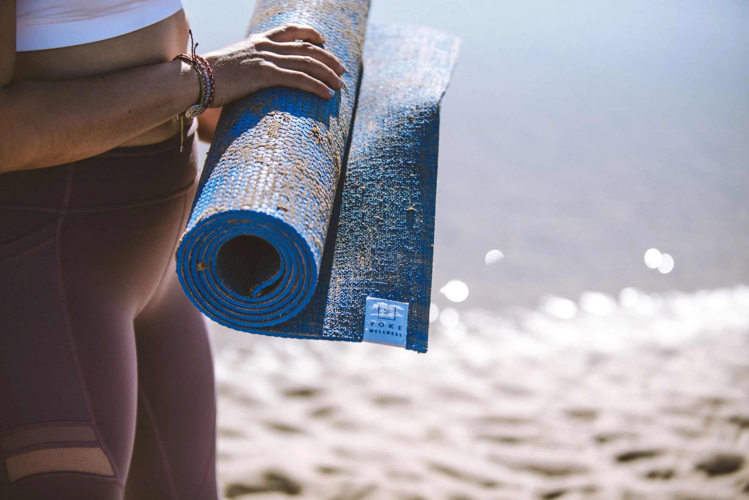 A person holding a rolled-up blue yoga mat at the beach with water and sand in the background.