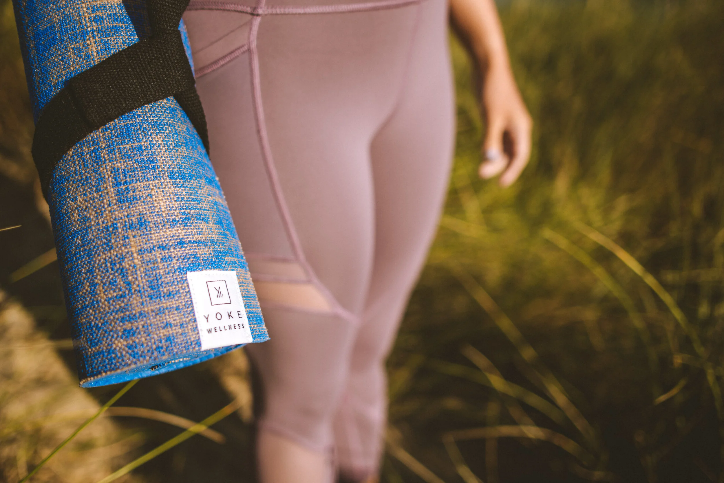 Close-up of a person's hand holding a blue yoga mat with a brand label, with the person wearing light pink leggings, outdoors in nature.