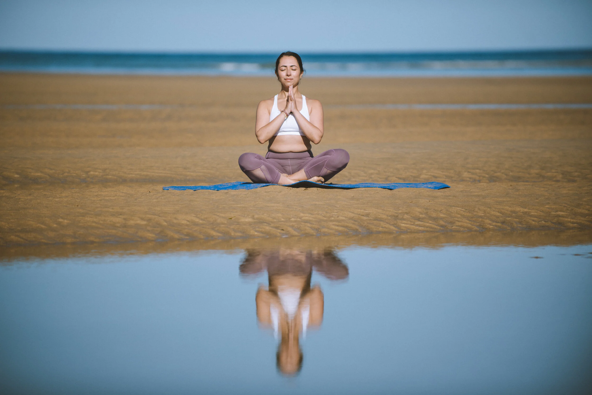 Woman practicing yoga on a blue mat on the beach with calm water reflecting her pose, ocean in the background