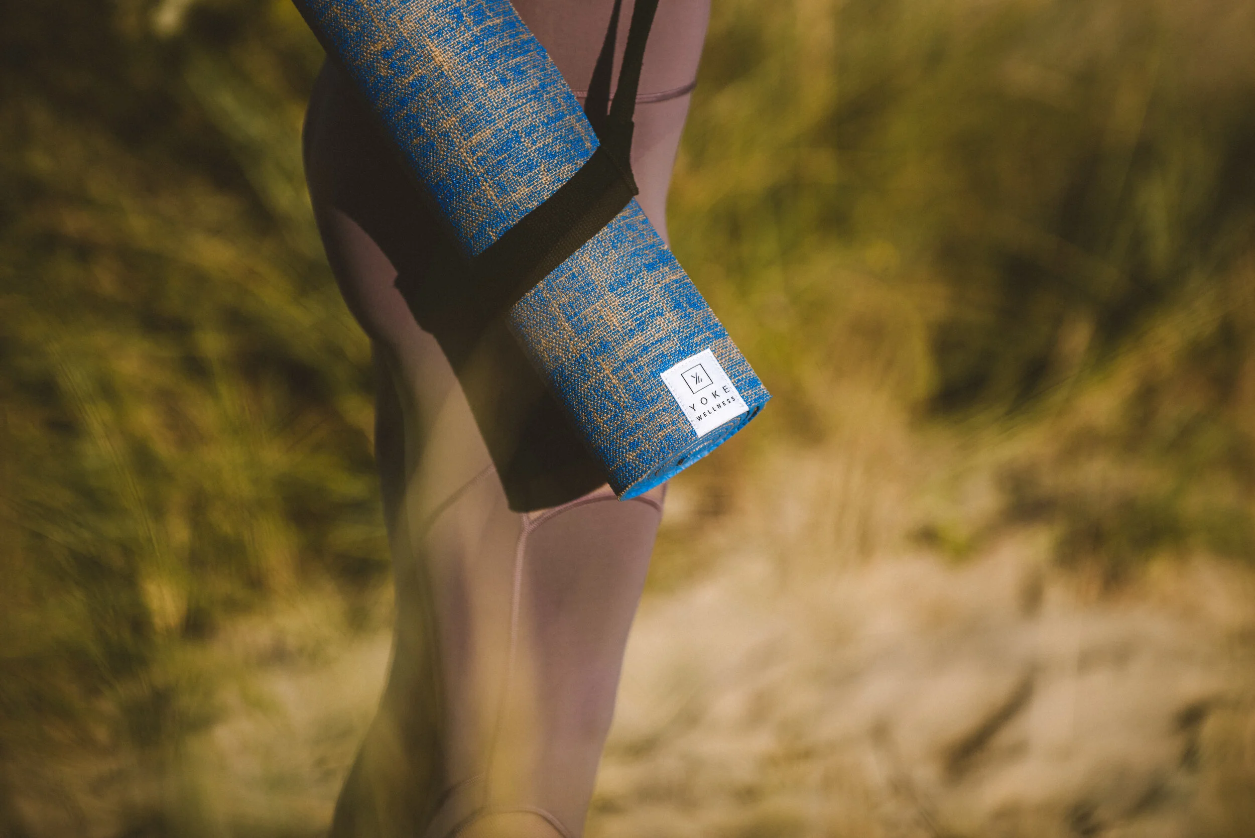 A person holds a blue yoga mat with a white label reading 'Yoke Wellness' while standing outdoors on a sandy trail surrounded by tall grass.