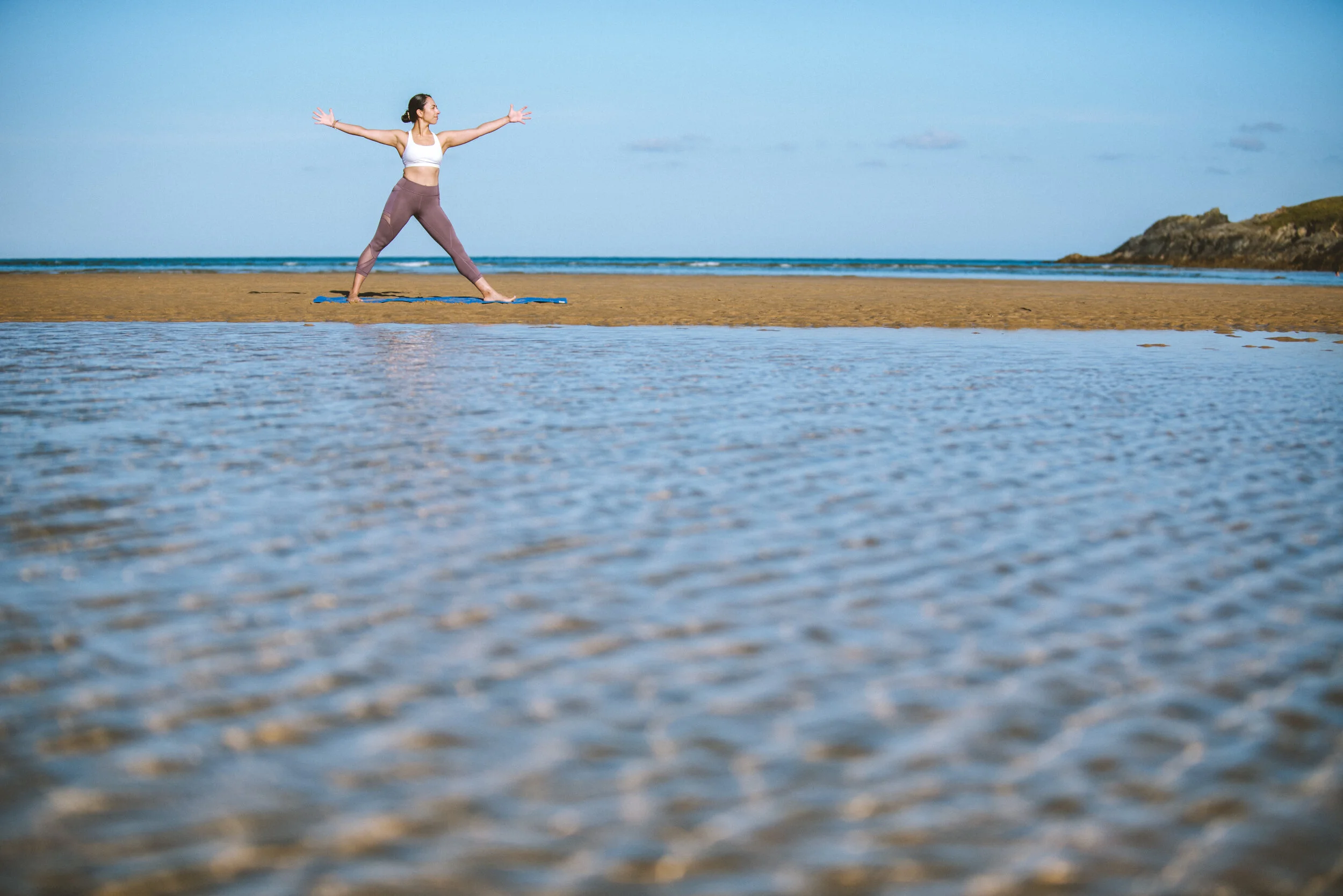 A woman practicing yoga on a yoga mat at the beach near the water with a scenic landscape in the background.