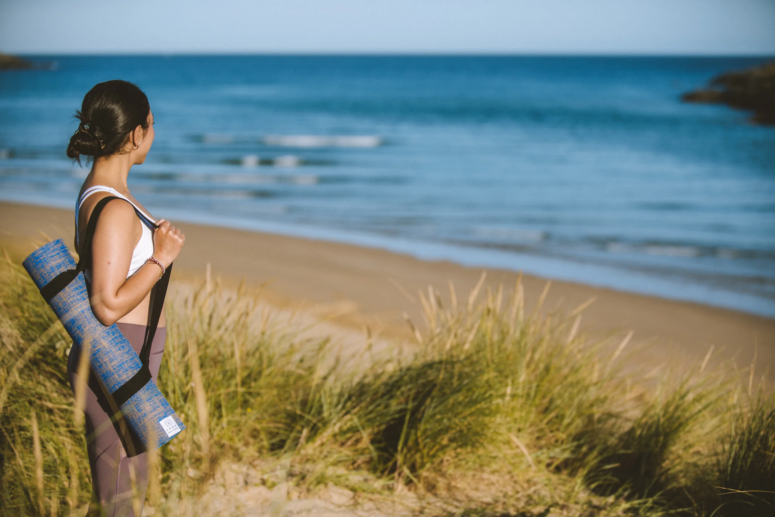 A woman stands on a sandy beach with grass, looking at the ocean waves, carrying a rolled-up yoga mat on her shoulder.