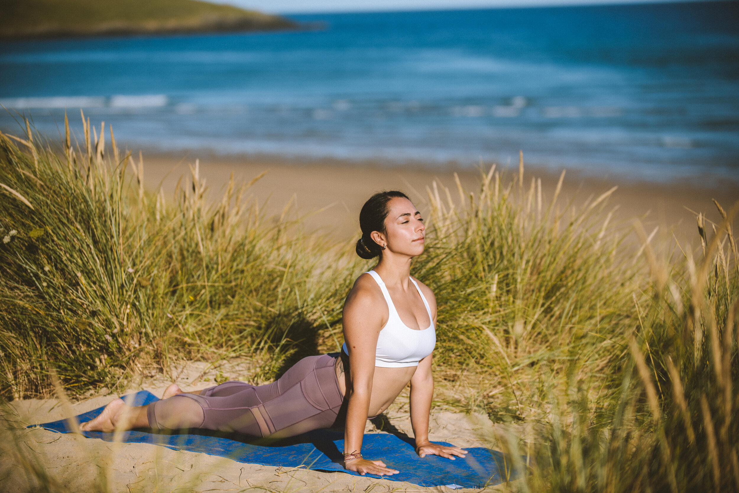 A woman practicing yoga outdoors on a sandy beach with grass, near the ocean, in a cobra pose with eyes closed.