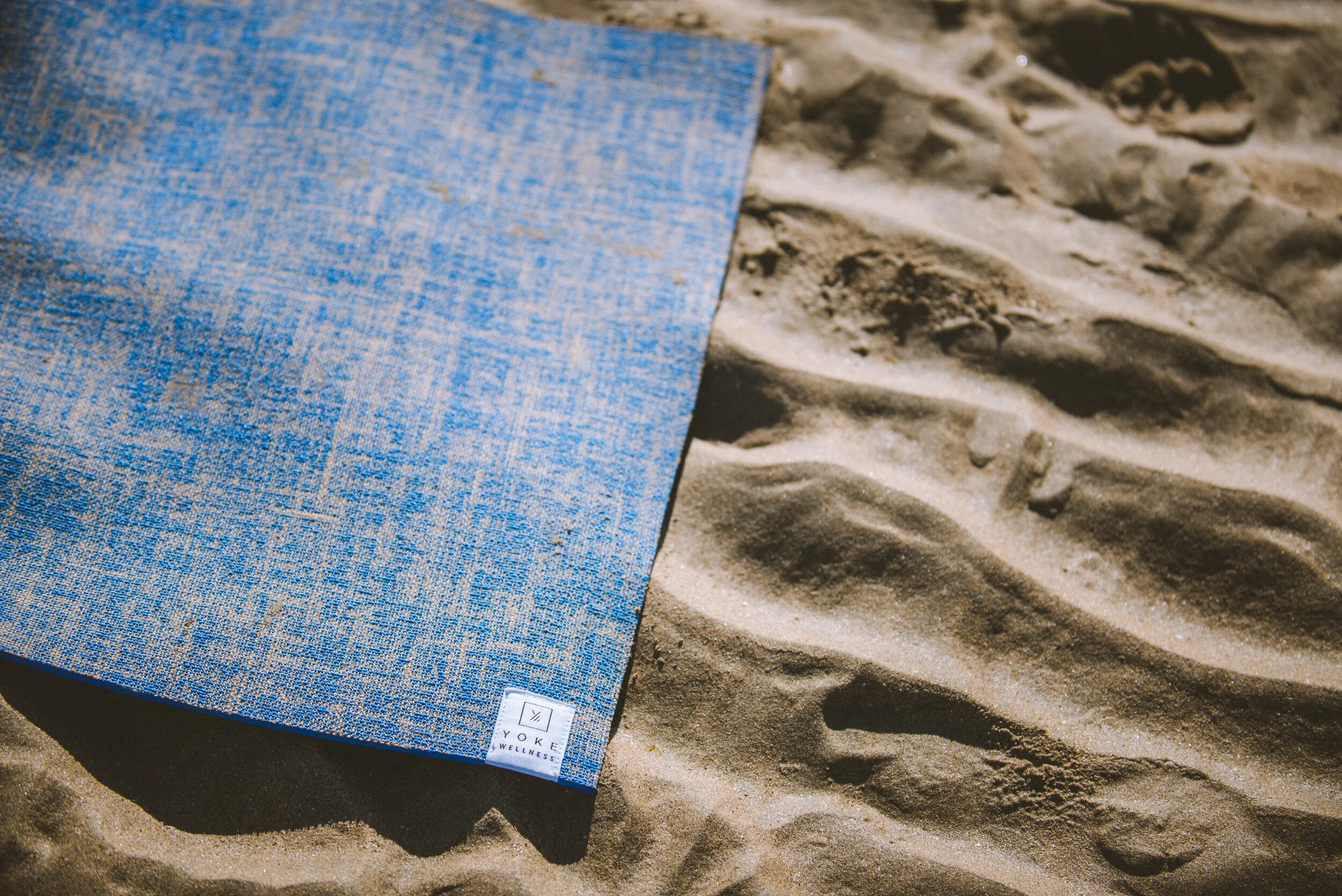A blue yoga mat with a woven pattern lying on sandy beach with footprints.