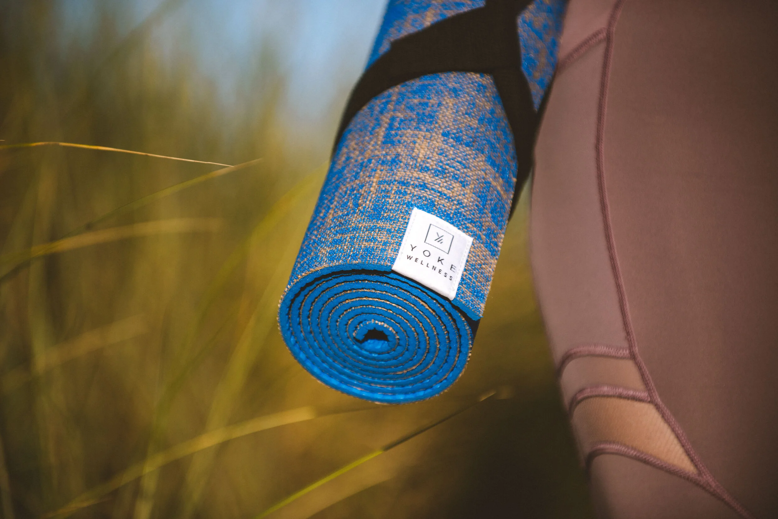 Close-up of a rolled blue and brown patterned yoga mat with a white label that reads 'VOKE WELLNESS,' held by a person wearing brown athletic leggings.