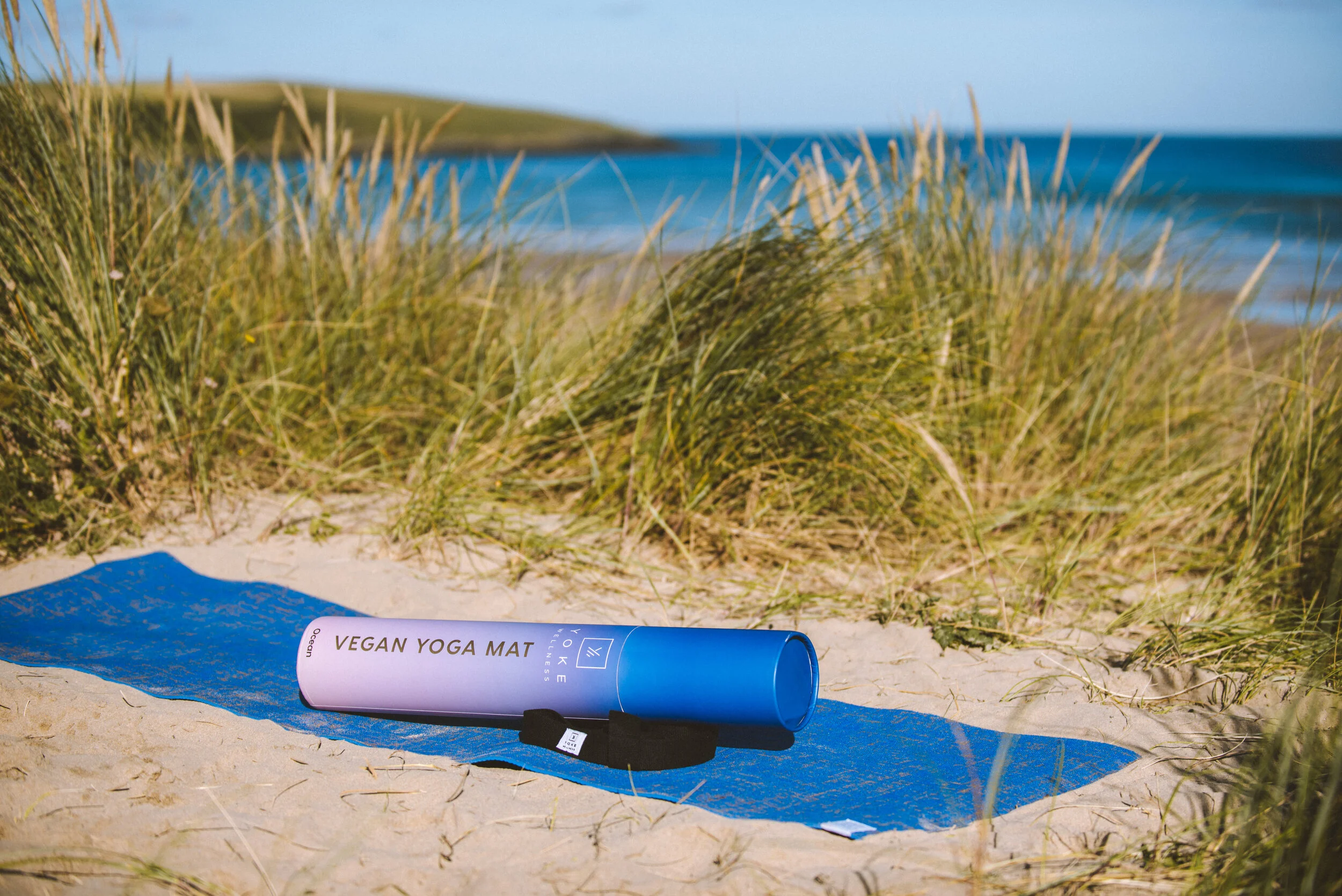 Vegan yoga mat rolled out on sand beach with grass and ocean in background.