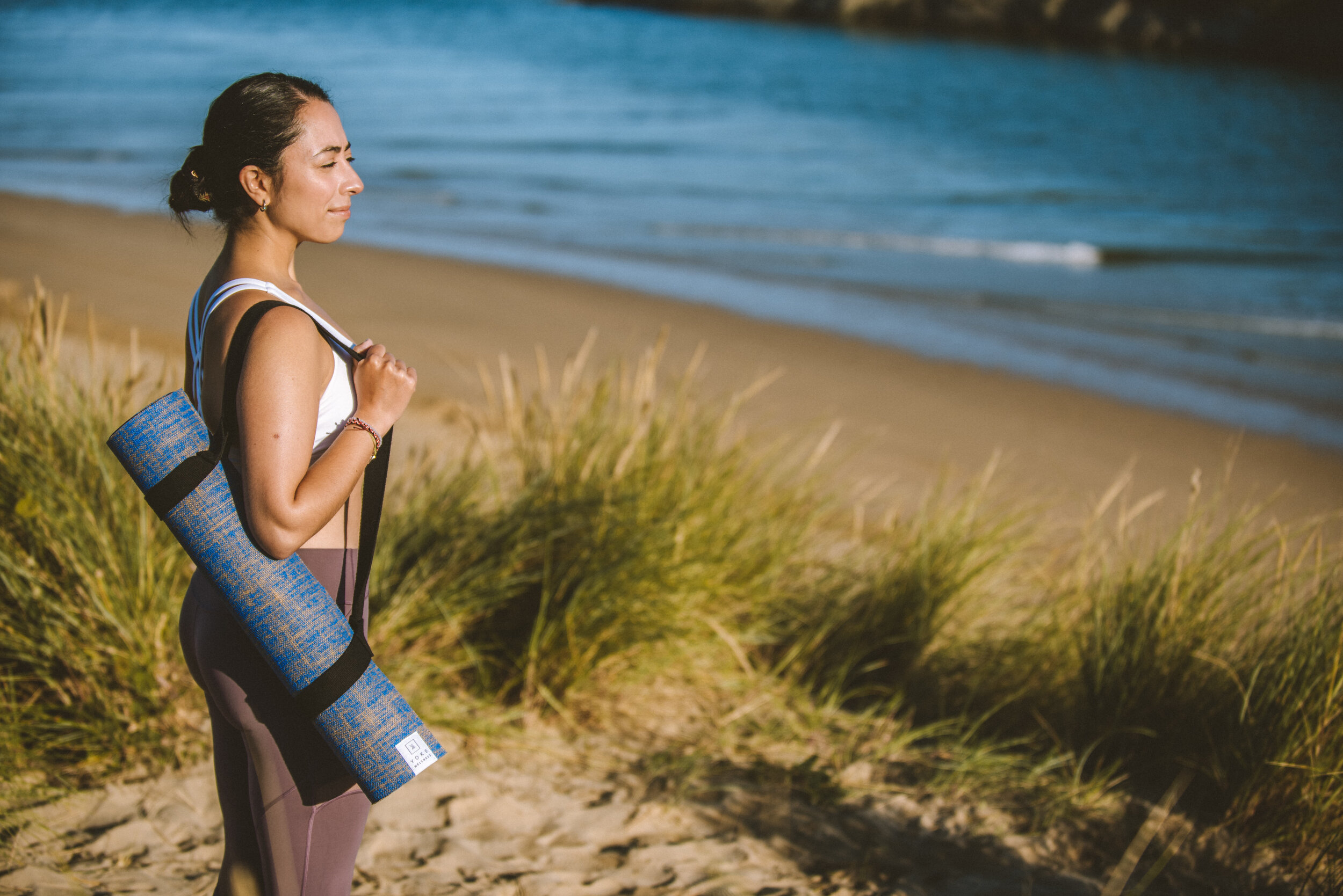 Woman carrying a yoga mat on her shoulder walking along a sandy beach with grass and the ocean in the background.