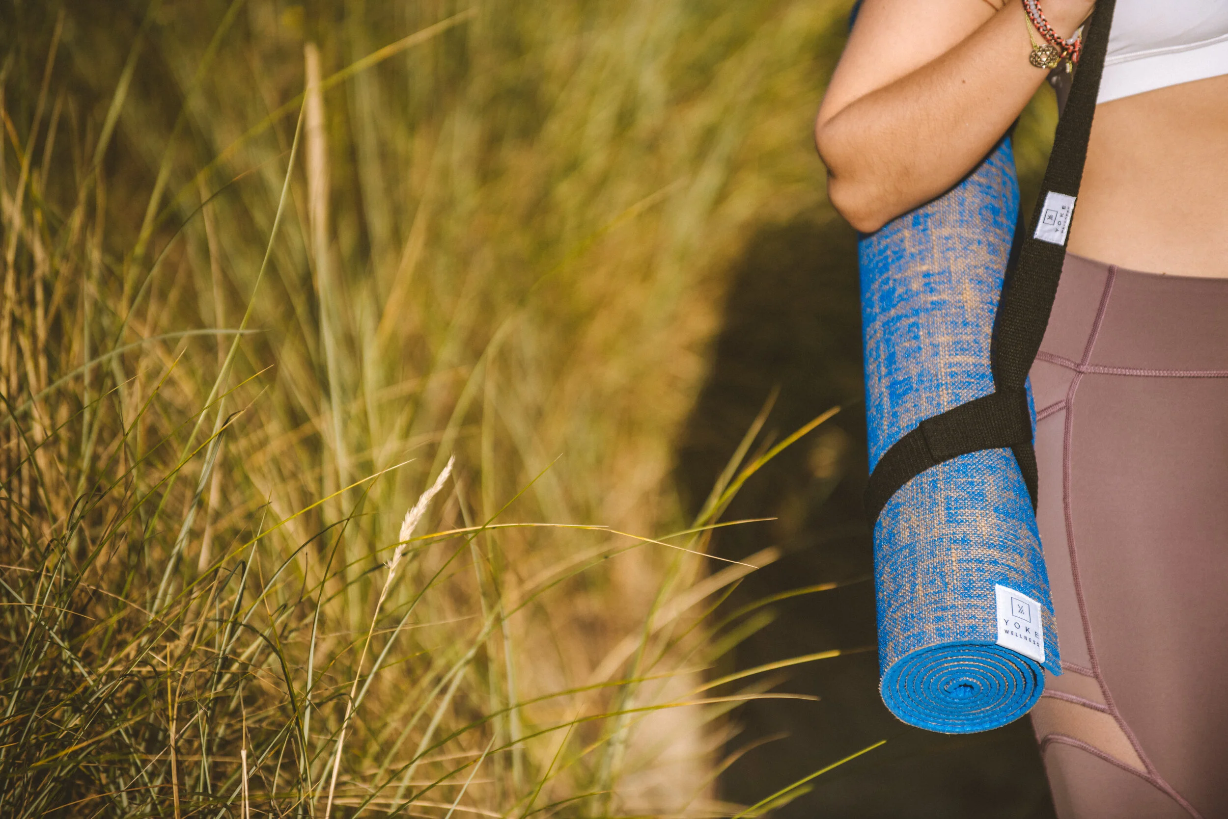 Person holding a rolled-up blue yoga mat on a hike in nature, with tall grass in the background.