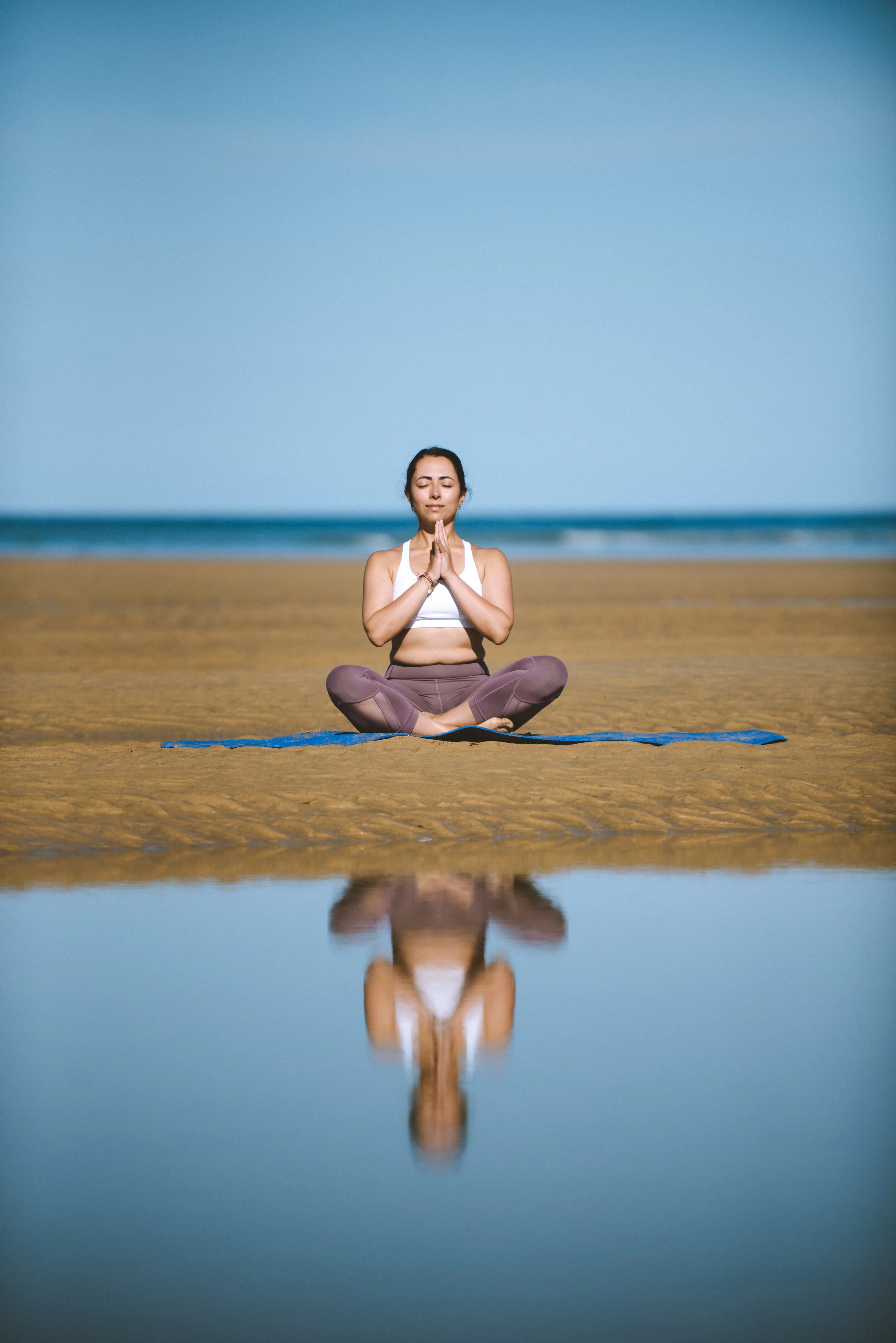 A woman practicing yoga on a beach, sitting cross-legged on a mat with hands in prayer position, reflecting in a small body of water.