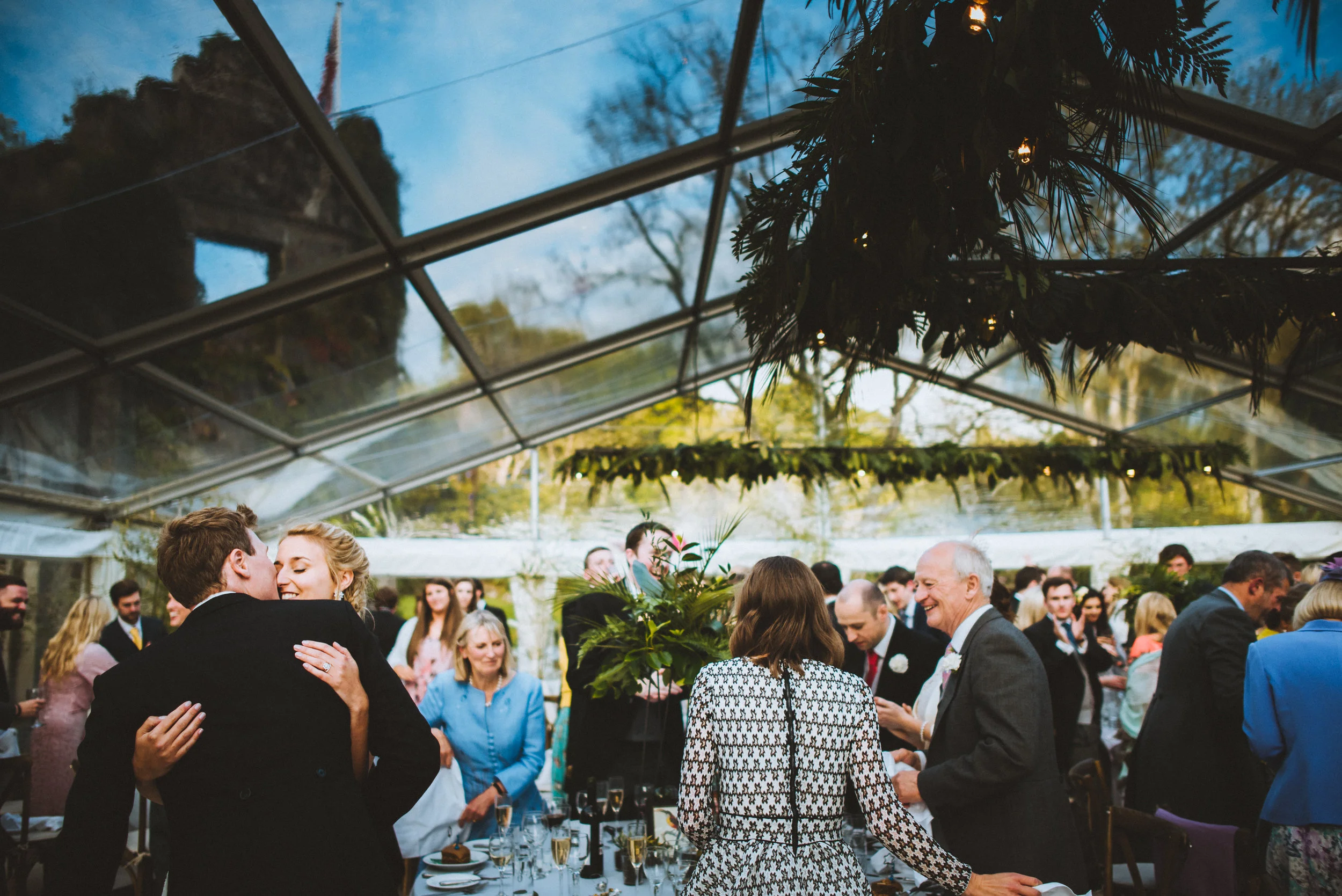 People celebrating at a wedding reception inside a decorated glass tent, with a couple dancing and guests around tables.