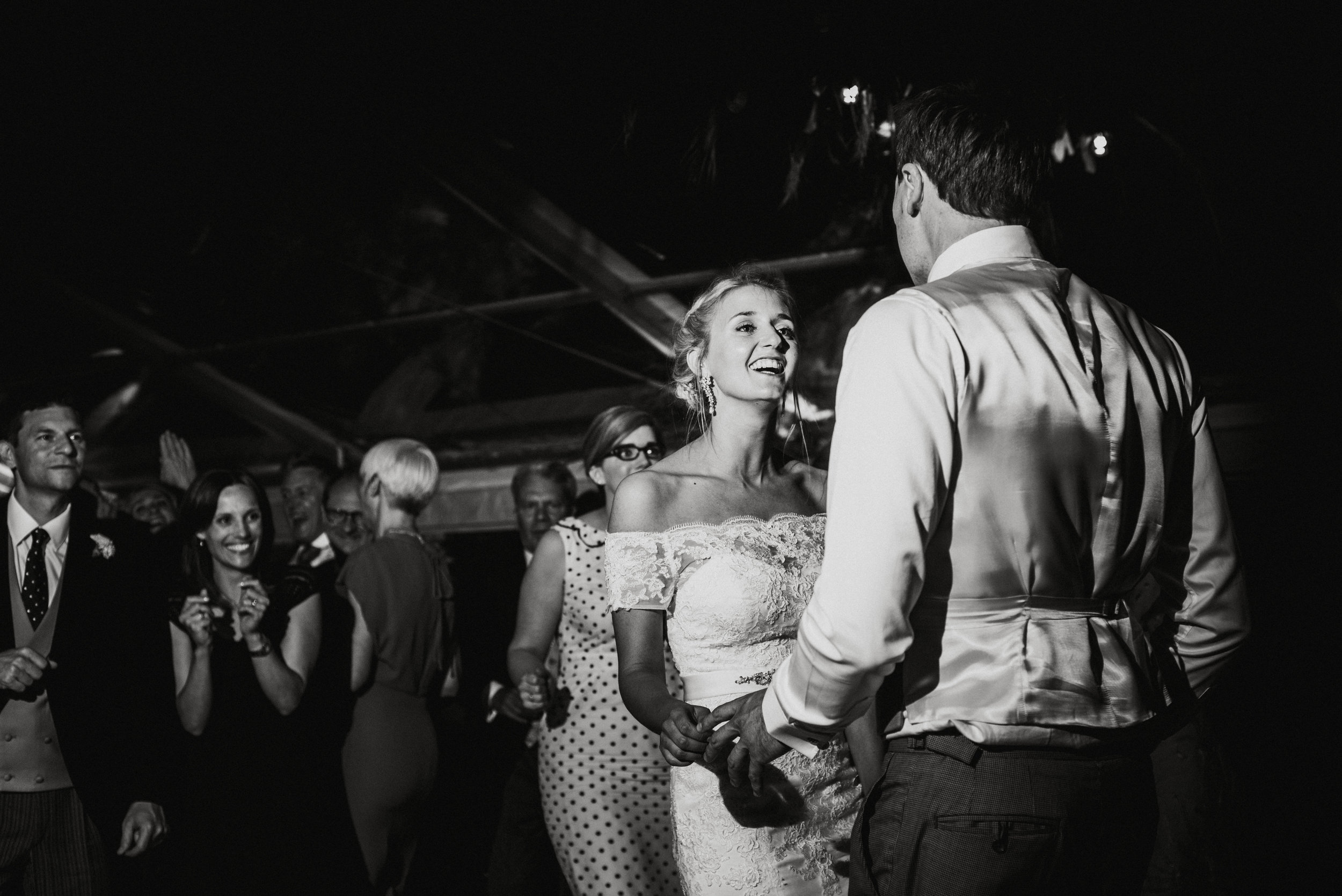 A bride and groom dancing together at their wedding reception, surrounded by their wedding guests, all in formal attire, in a black-and-white photo.
