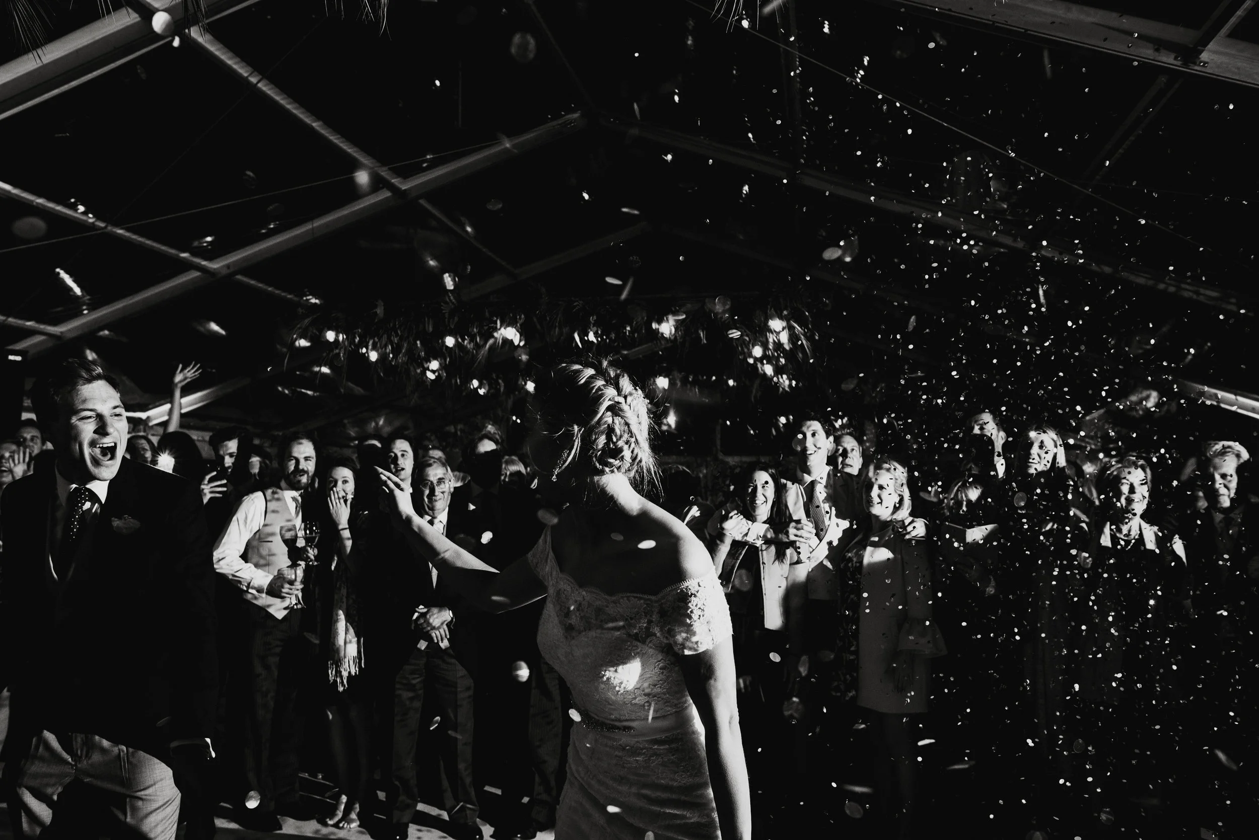 Black and white photo of a wedding reception with a bride in a lace dress dancing and confetti falling, surrounded by guests smiling and cheering.