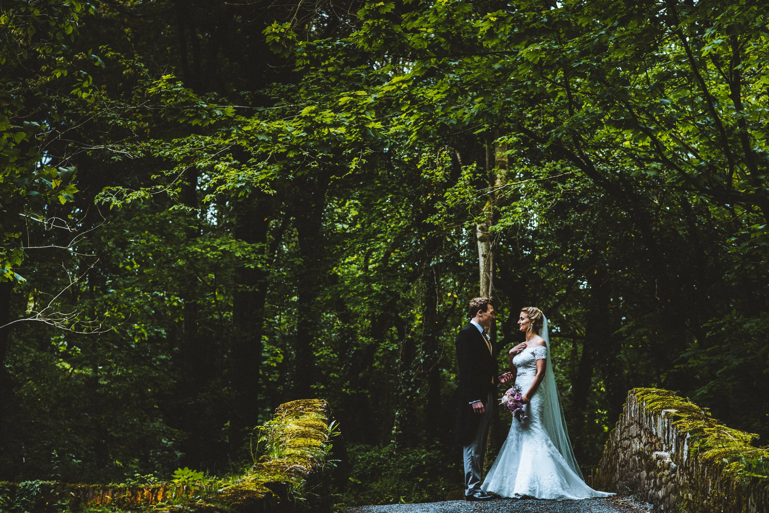 Wedding photo of A bride and groom stand on a moss-covered bridge in a dense, green forest, gazing at each other on their wedding day.
