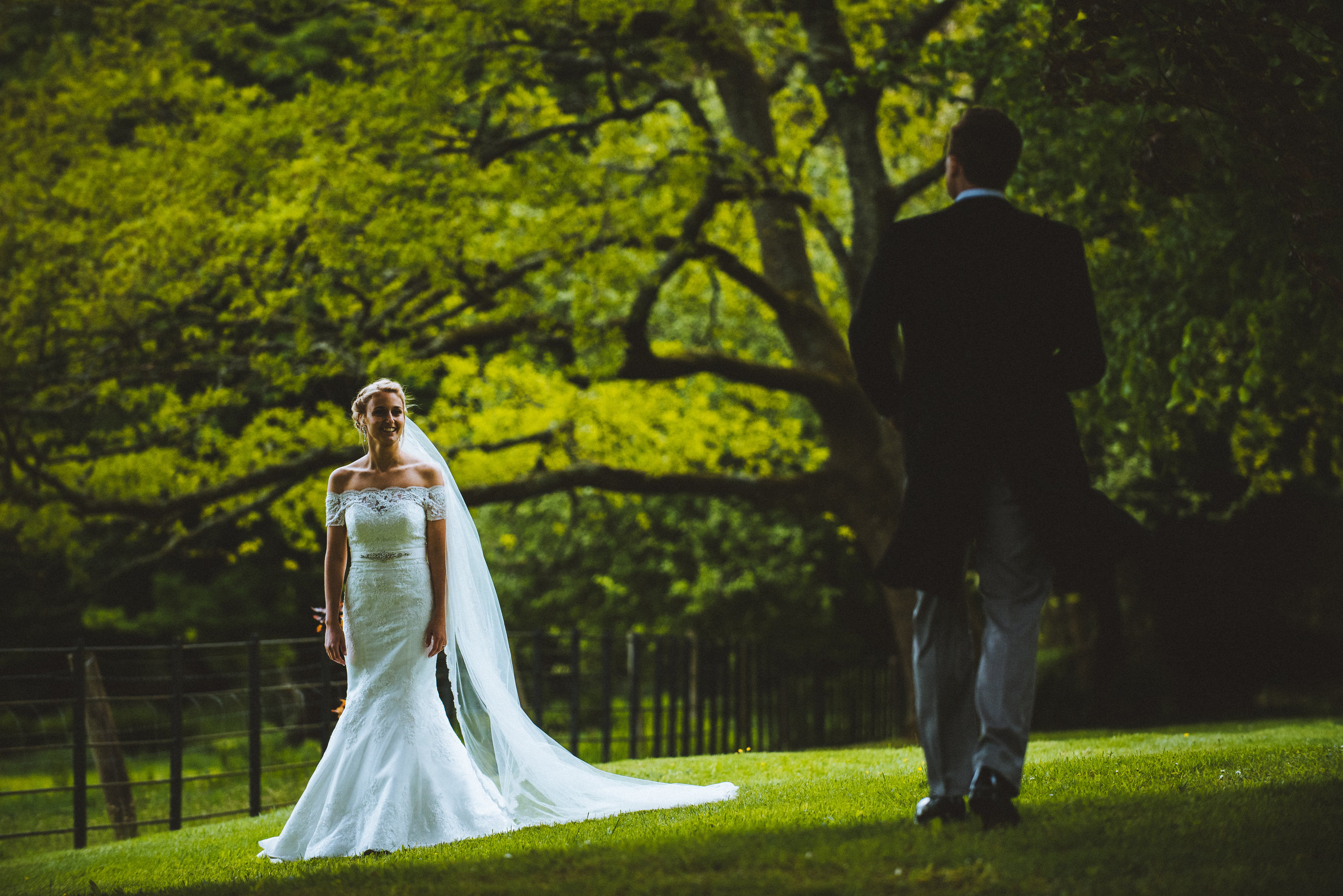 A bride in a white wedding dress stands on green grass in a park, smiling at a man in a suit walking towards her, with trees and a fence in the background.