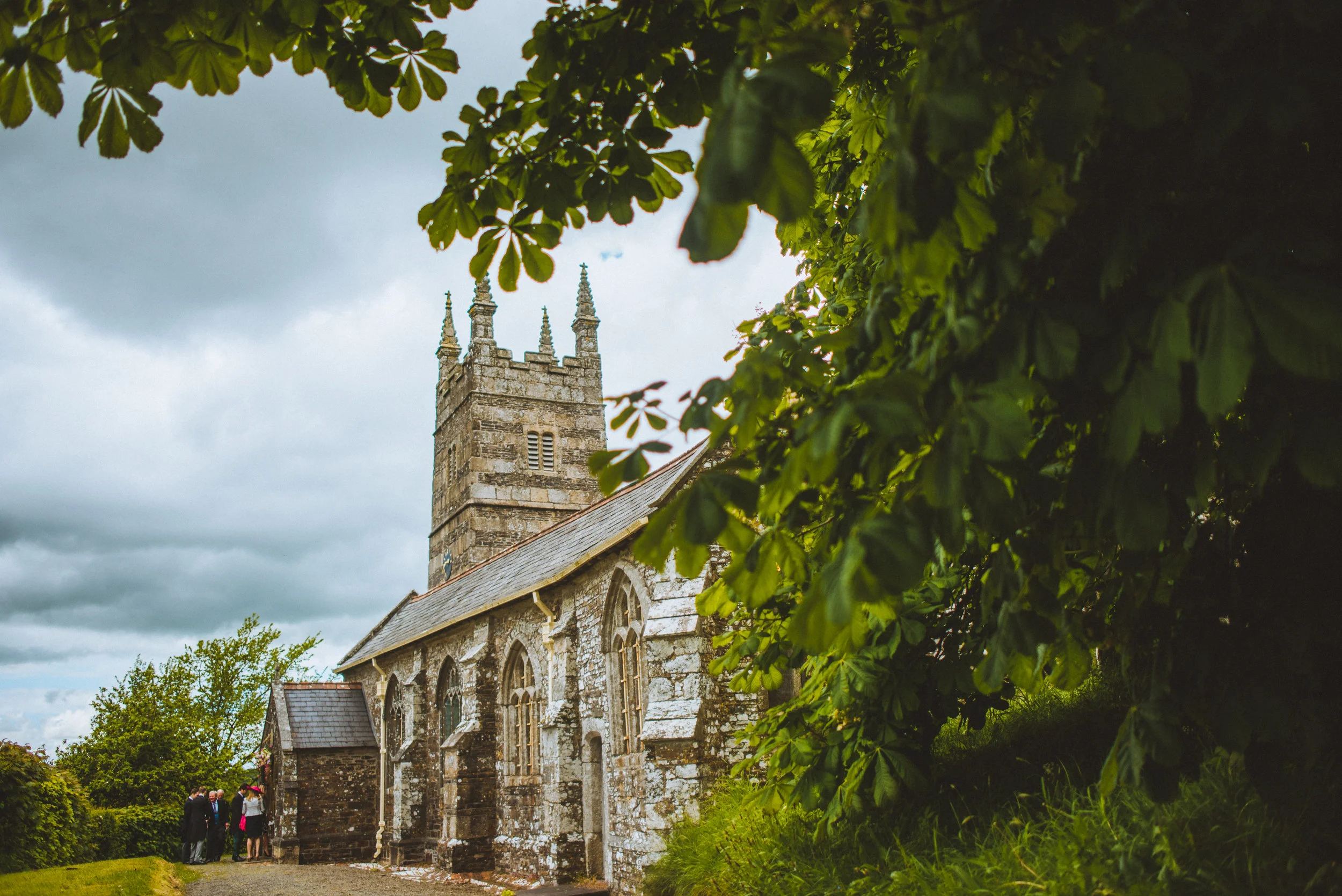 Isles of scilly photography. A historic stone church with a tall steeple and Gothic-style windows, surrounded by greenery, with a group of people gathered outside under cloudy skies.